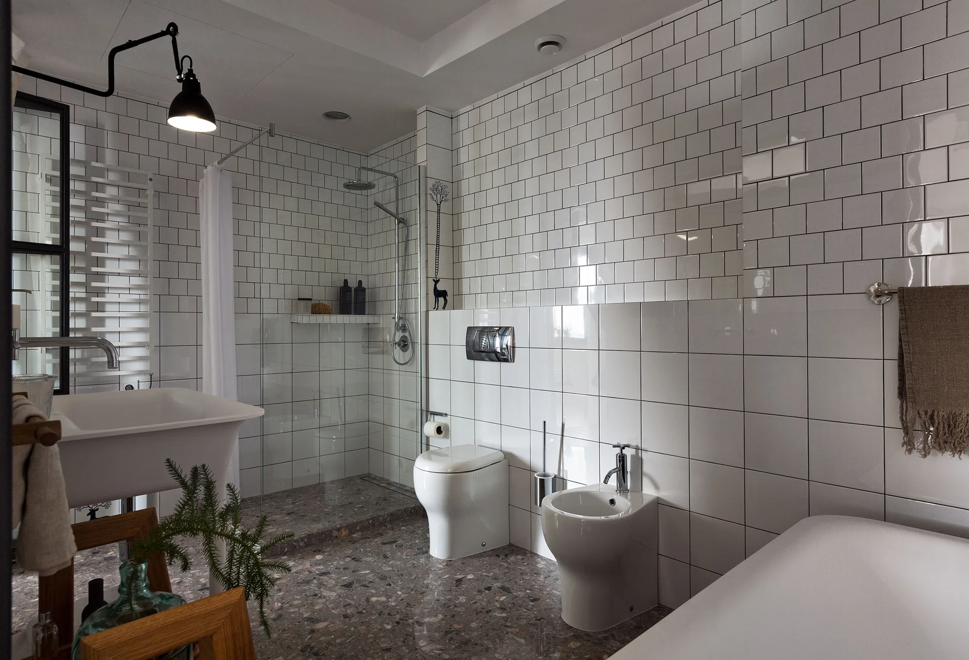 A modern bathroom features white tiled walls, a speckled grey terrazzo floor, a wall-hung toilet, and a walk-in glass shower.