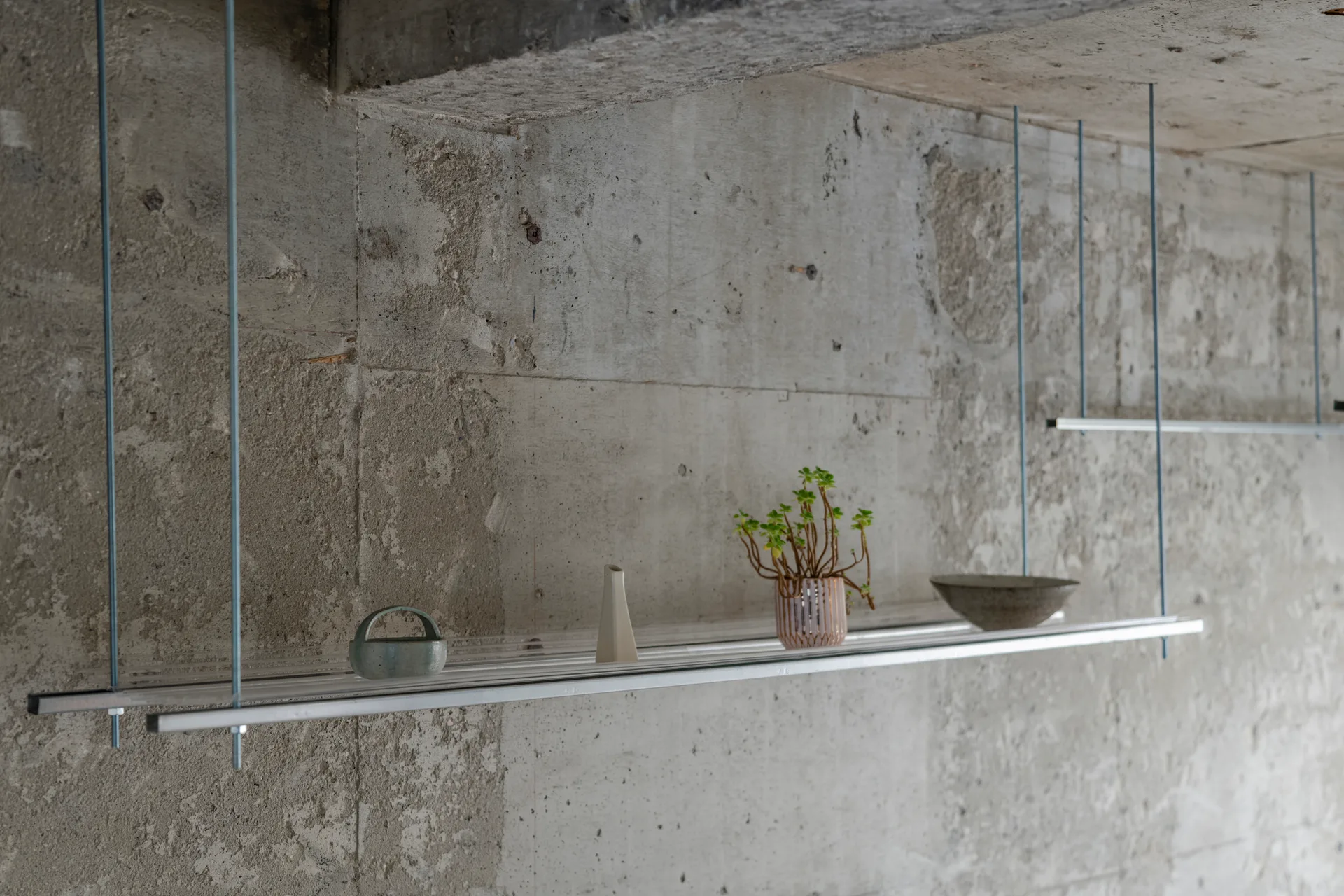 A thin metal shelf suspended by rods from a raw concrete ceiling hangs against a textured concrete wall, holding small ceramic decor items.