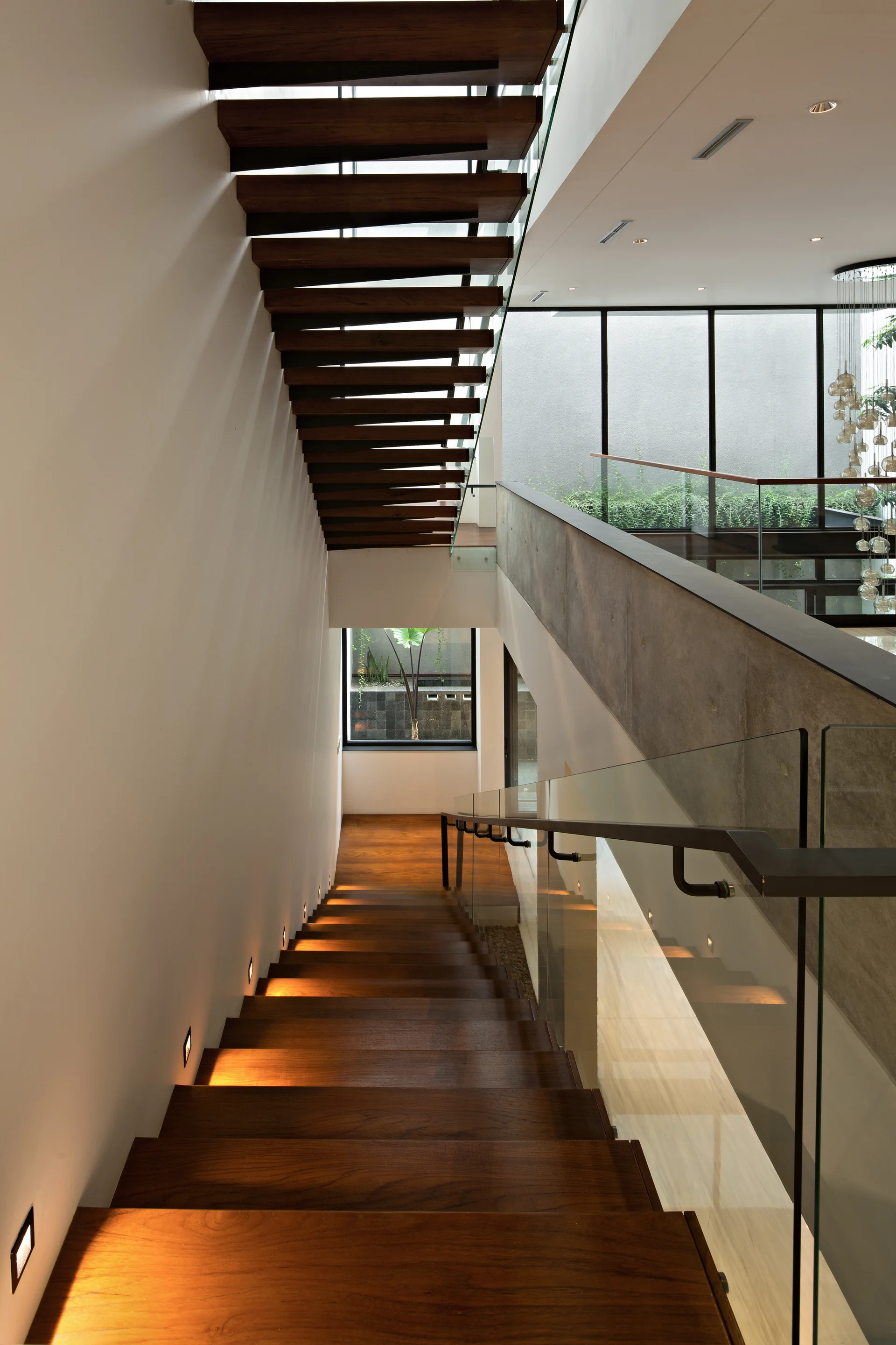 A modern floating wood staircase with clear glass handrails and recessed square wall lights leading down to a bright landing.