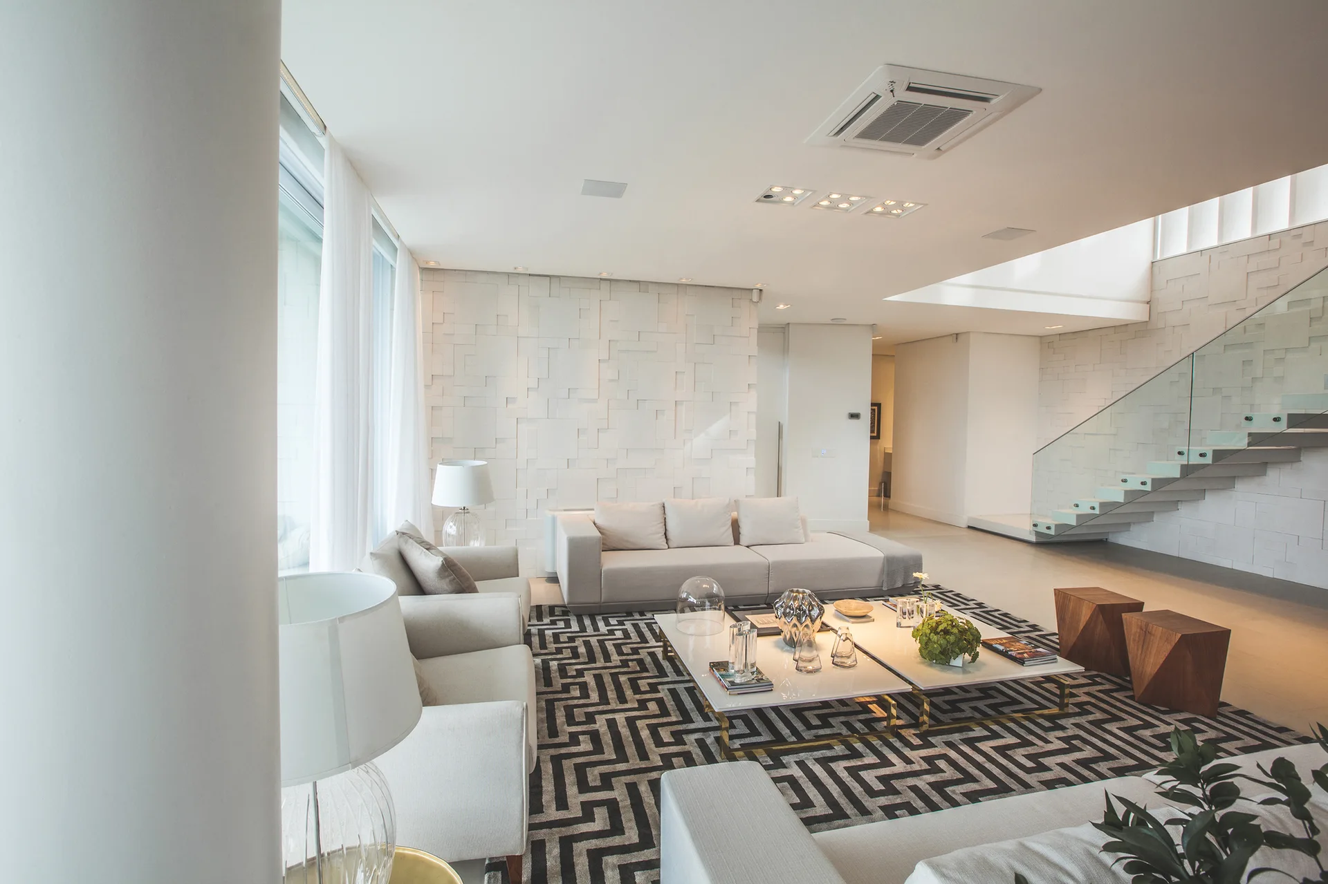 A bright living room features a large beige modular sofa, a bold geometric patterned rug, and a floating staircase with glass railings in the background.