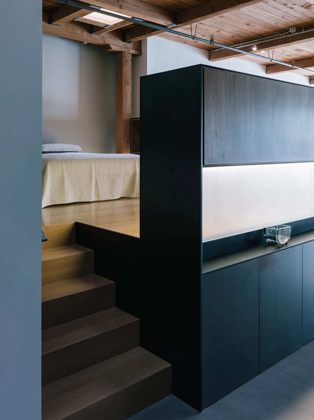 Wooden stairs lead up to a raised bedroom platform beside a matte black cabinet unit with an integrated backlit glass panel and exposed ceiling beams.