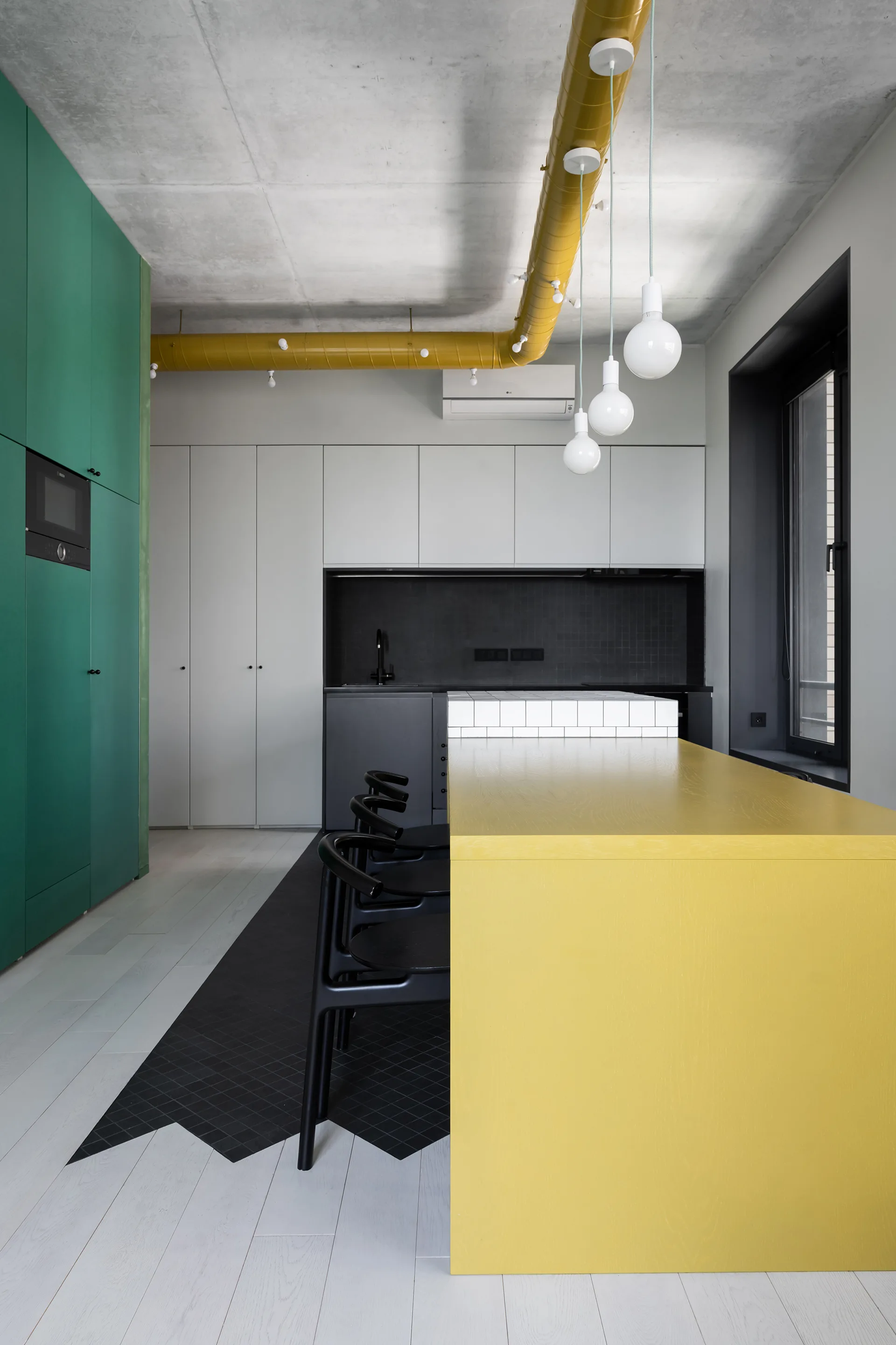 A modern kitchen with a bold yellow island, black counter stools, matte green cabinets, and a yellow painted ceiling duct against concrete.