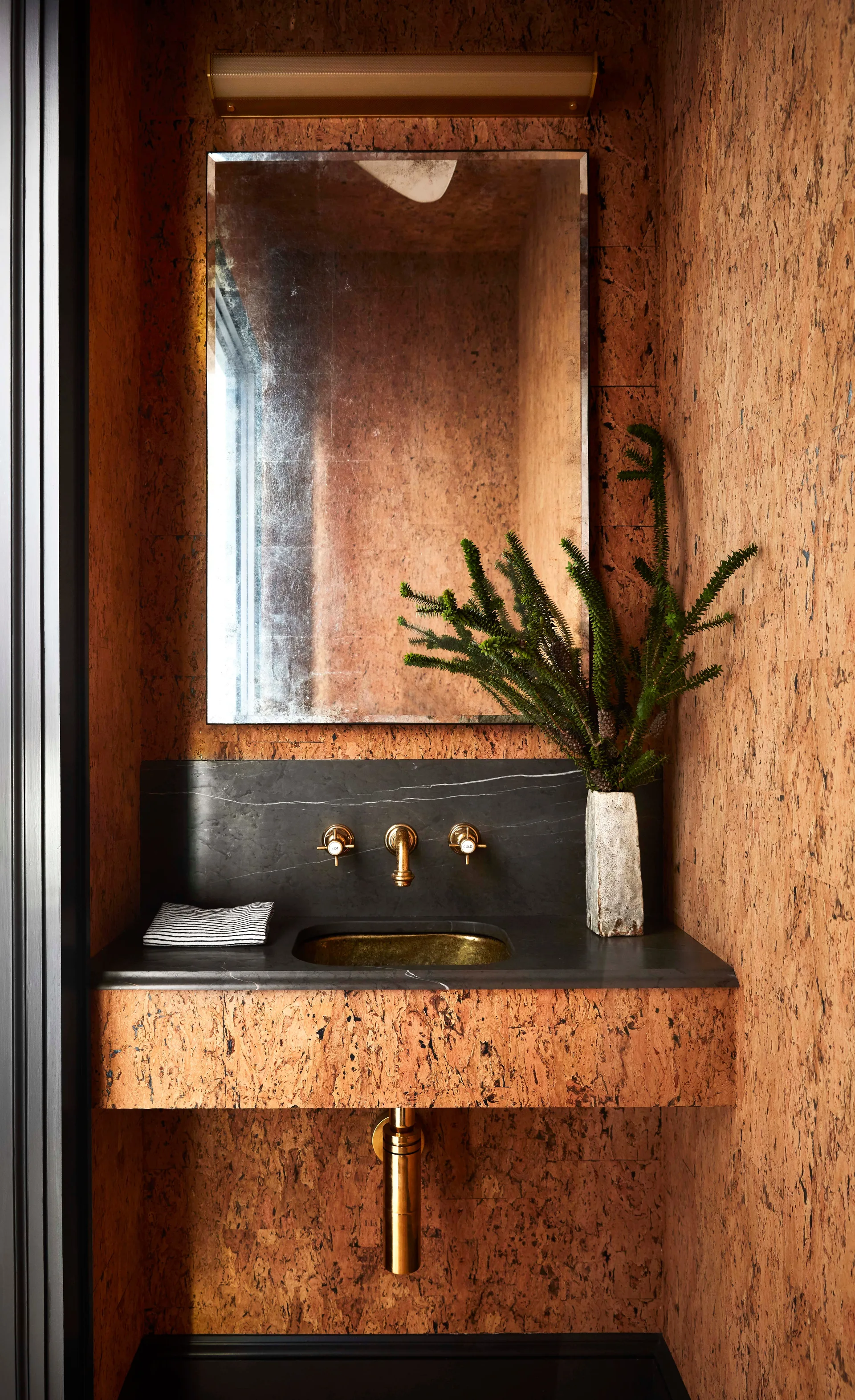 A compact powder room features textured cork wall panels, a dark stone vanity with a brass sink, and a matching wall-mounted brass faucet.