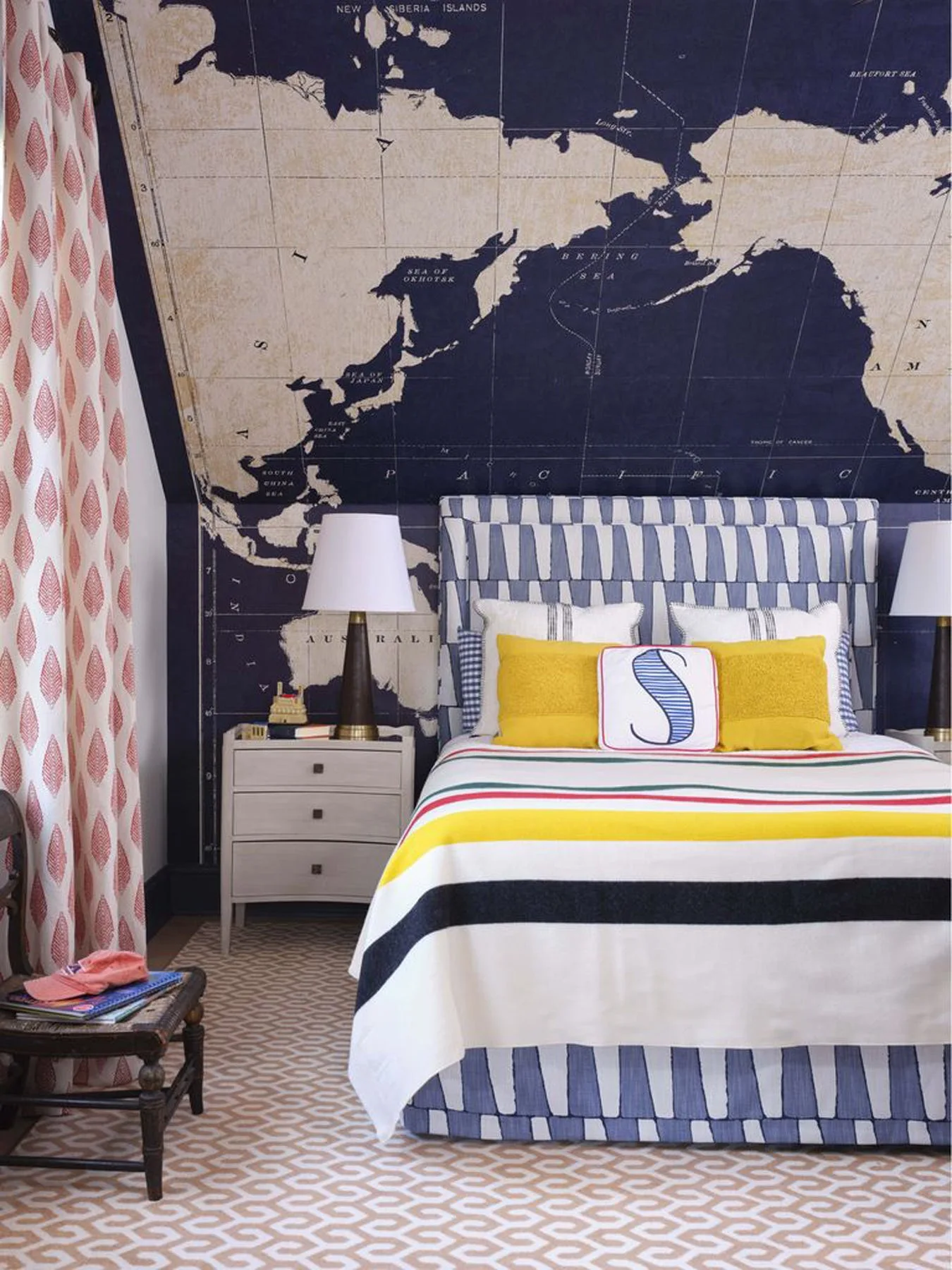 A bedroom features a navy blue world map wall mural behind a striped upholstered bed with colorful pillows and a matching white nightstand.