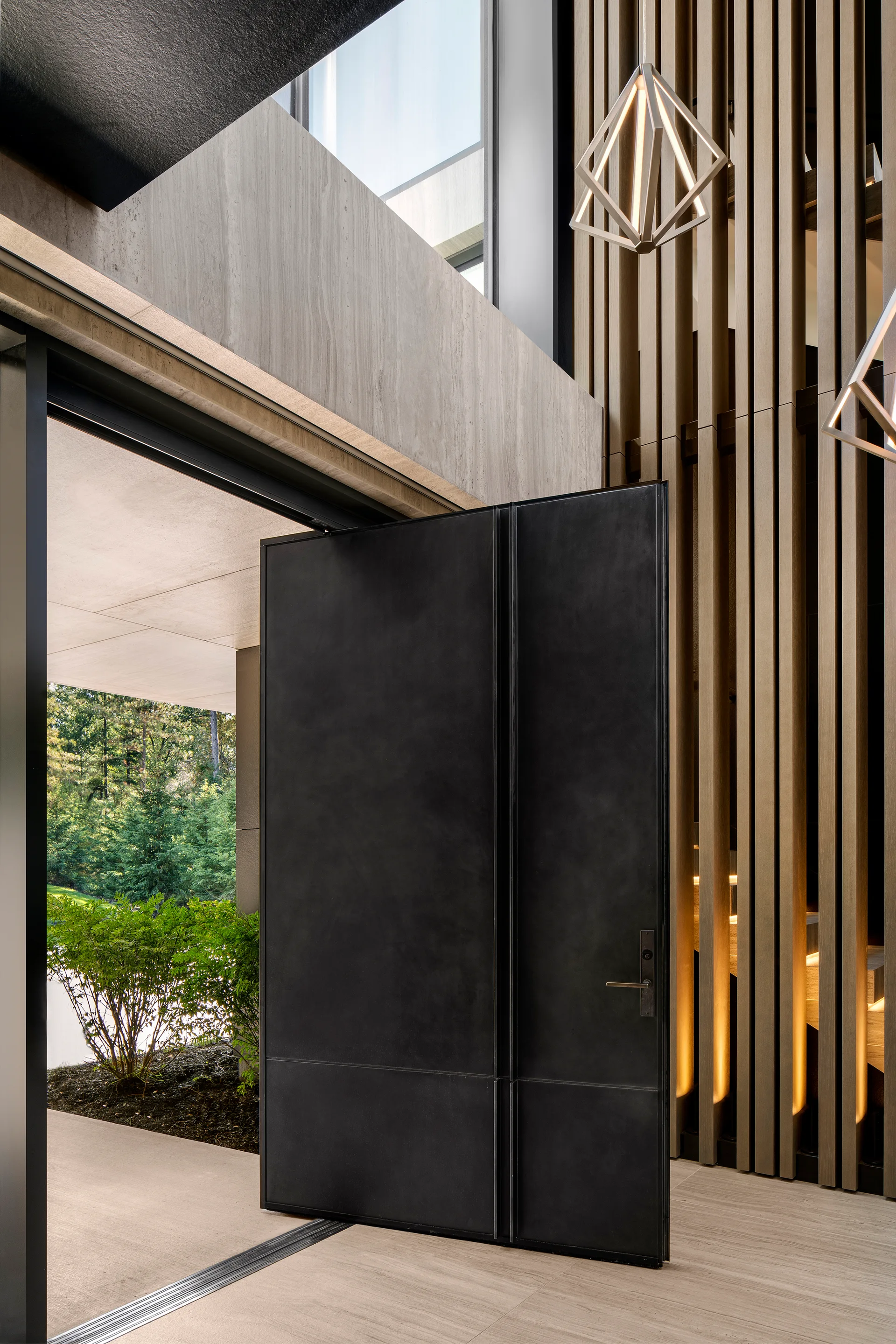 A tall matte black pivot door stands open next to a vertical wood slat wall with integrated accent lighting and a geometric pendant.