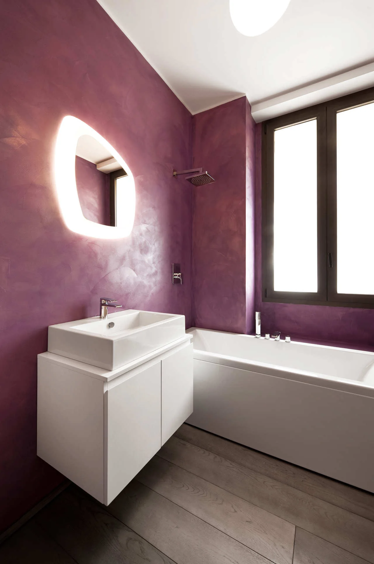A modern bathroom features textured purple plaster walls, a floating white vanity with a vessel sink, a backlit mirror, and a white bathtub.