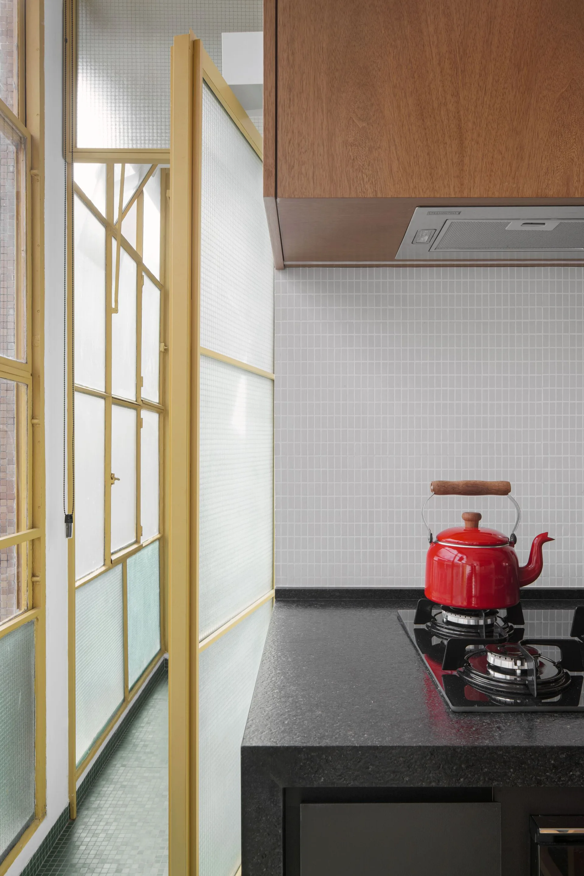 A red kettle sits on a black gas cooktop next to a white grid tile backsplash and a yellow metal window frame.