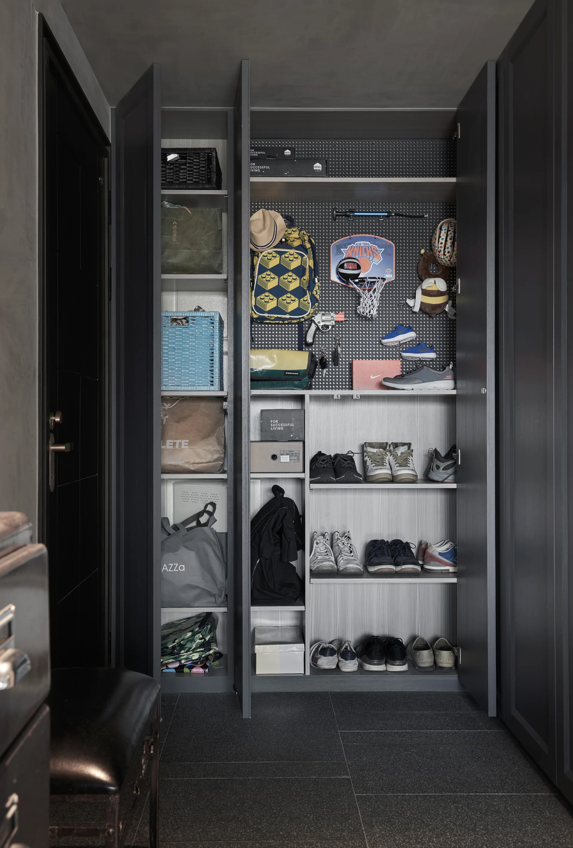 A dark gray built-in wardrobe with open doors reveals shelves lined with shoes and a perforated pegboard holding bags and accessories.