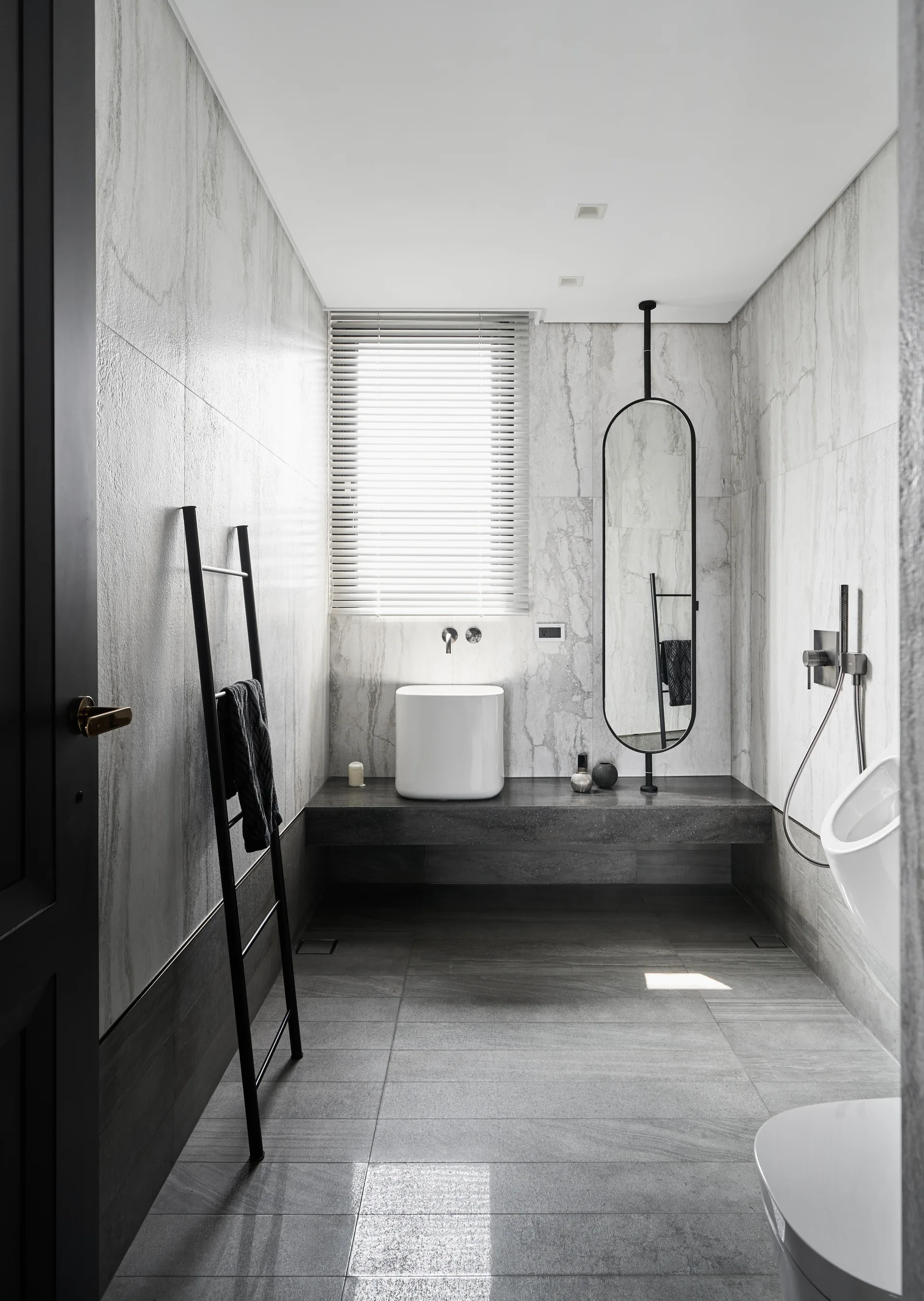 A minimalist bathroom with grey marble walls, a low stone vanity, a white vessel sink, a tall oval mirror, and a black leaning towel ladder.