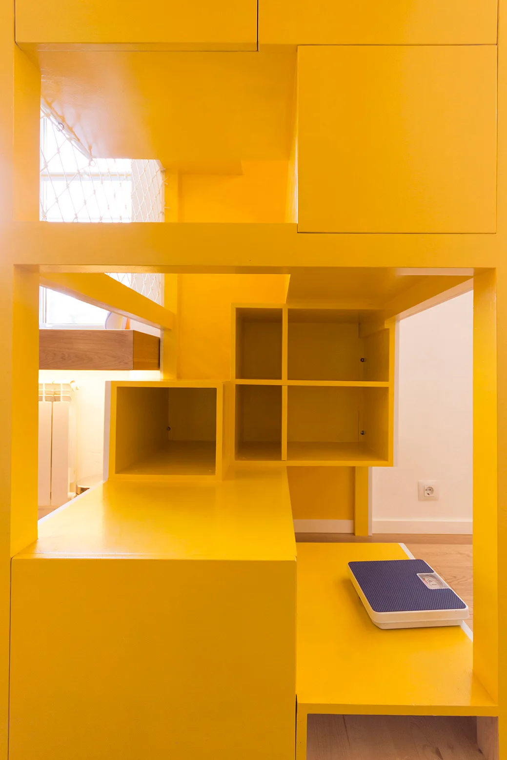 A bright yellow modular built-in shelving unit with geometric compartments sits next to a light wood floor with a blue scale.
