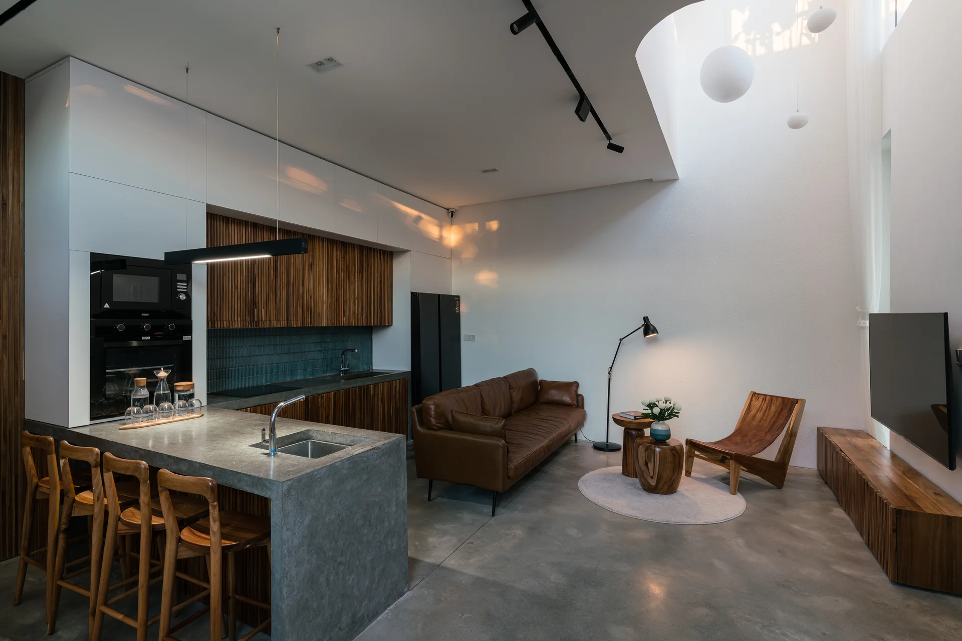 An open-plan living room and kitchen featuring a concrete island with barstools, a brown leather sofa, and vertical slatted wood cabinets.