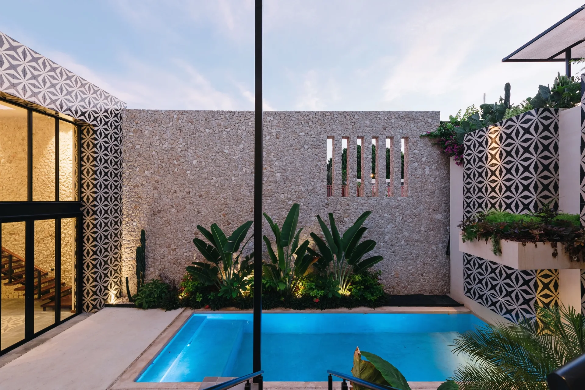 An outdoor courtyard swimming pool surrounded by a tall textured stone wall, tropical plants, and black-and-white patterned tiles.