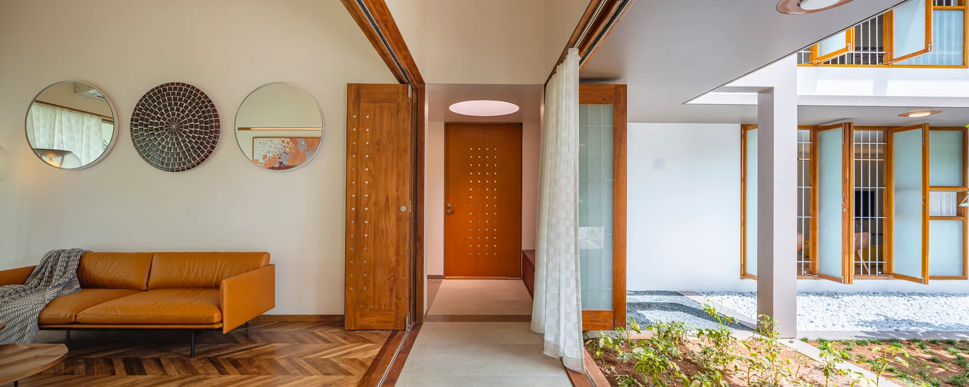 A bright room features a tan leather sofa, herringbone wood flooring, circular wall mirrors, and an adjacent courtyard filled with white pebbles.