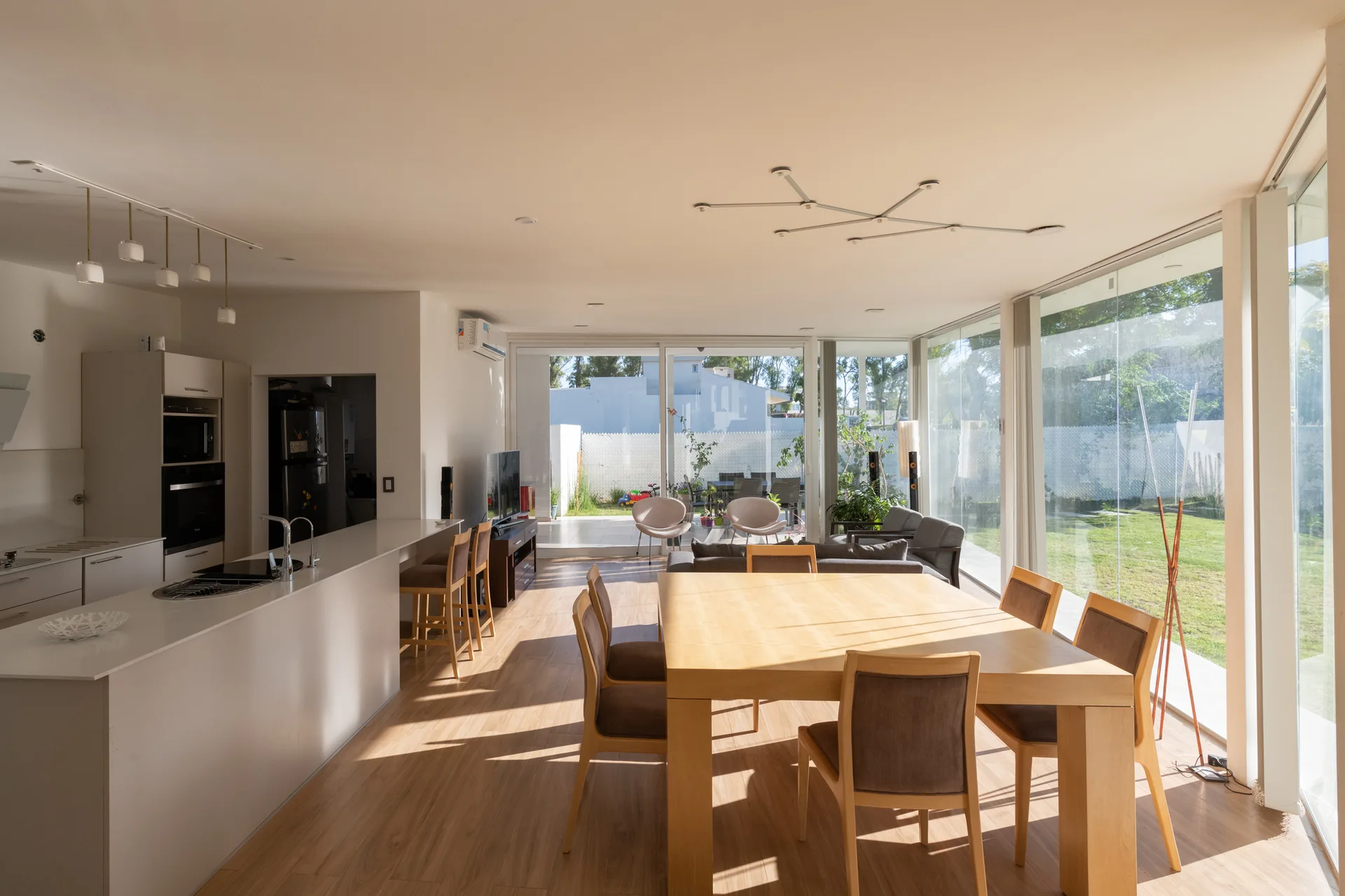 A light-wood dining table and a white kitchen island sit in an open-plan room with floor-to-ceiling glass walls showing a garden outside.