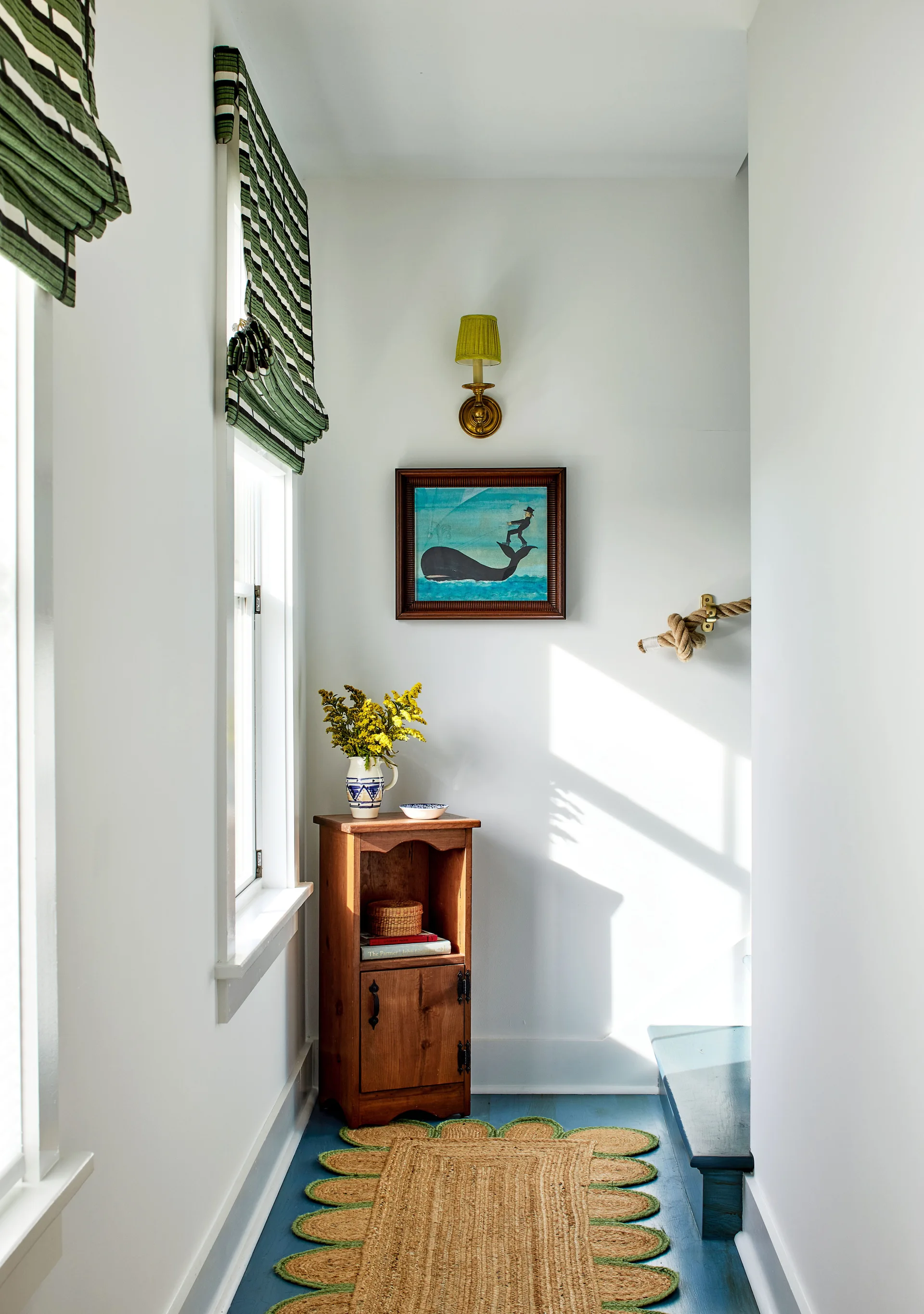 A narrow white hallway featuring a wooden corner cabinet, green striped roman shades, and a scalloped jute rug on painted blue floors.