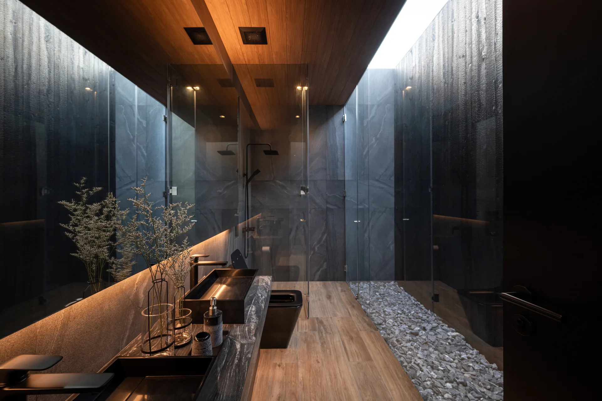 A modern bathroom with a dark stone vanity, double black vessel sinks, a warm wood ceiling, and a glass-enclosed shower with a white pebble floor.