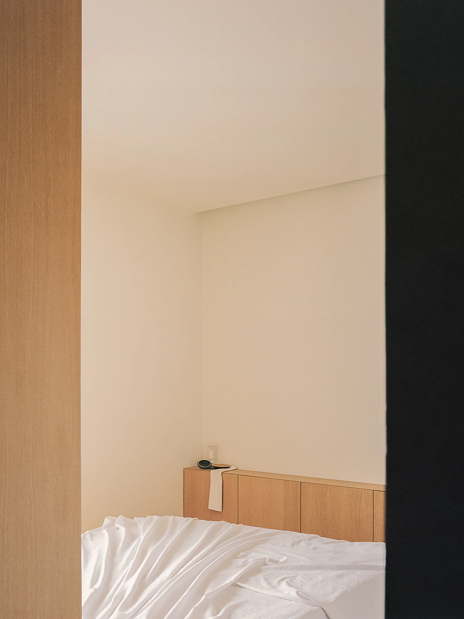 A minimalist bedroom viewed through a doorway, showing a bed with white sheets and a low built-in wood-toned cabinet acting as a headboard.