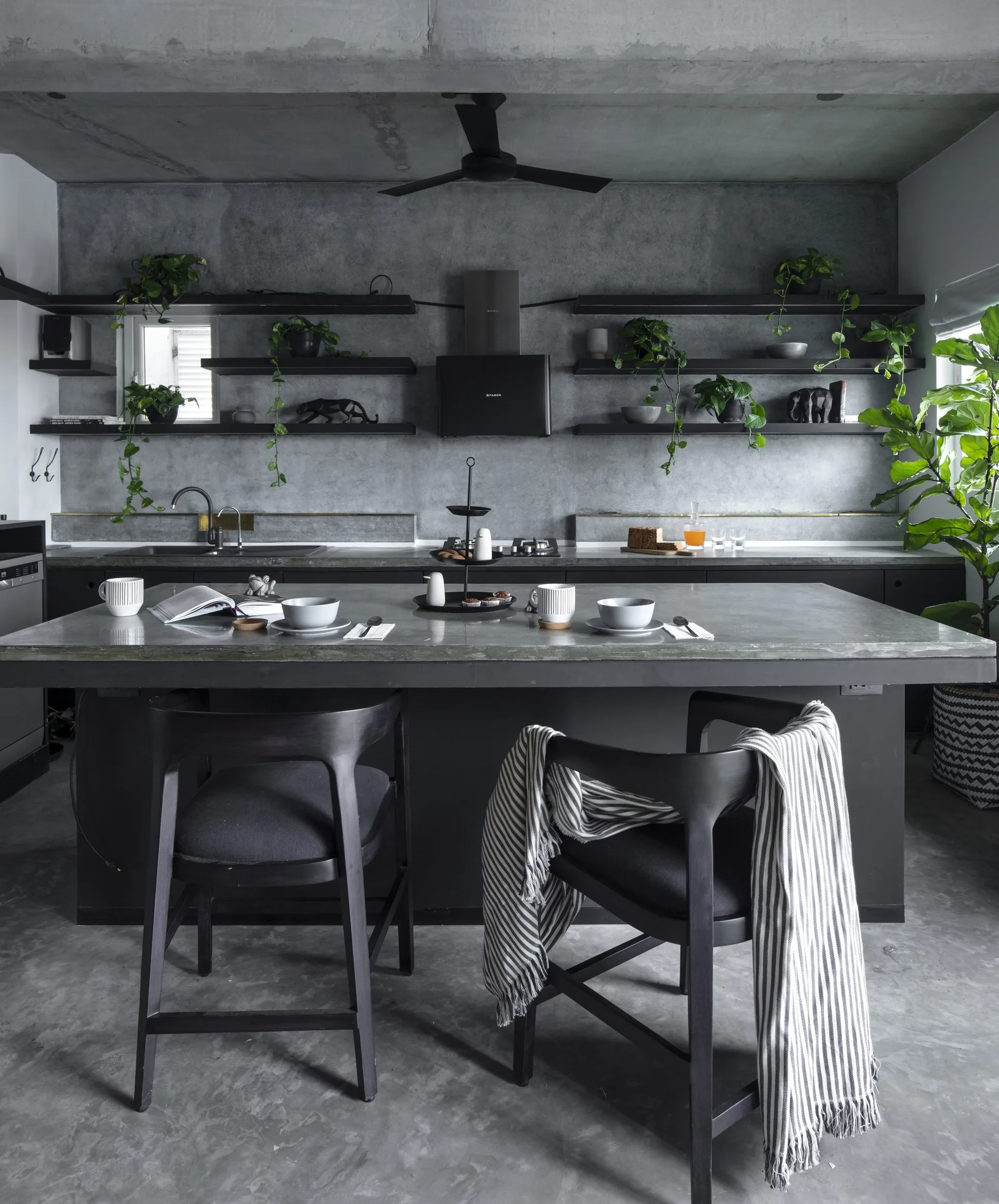 A modern industrial kitchen features a concrete island with black bar stools, set against a concrete wall with black open shelves and trailing plants.