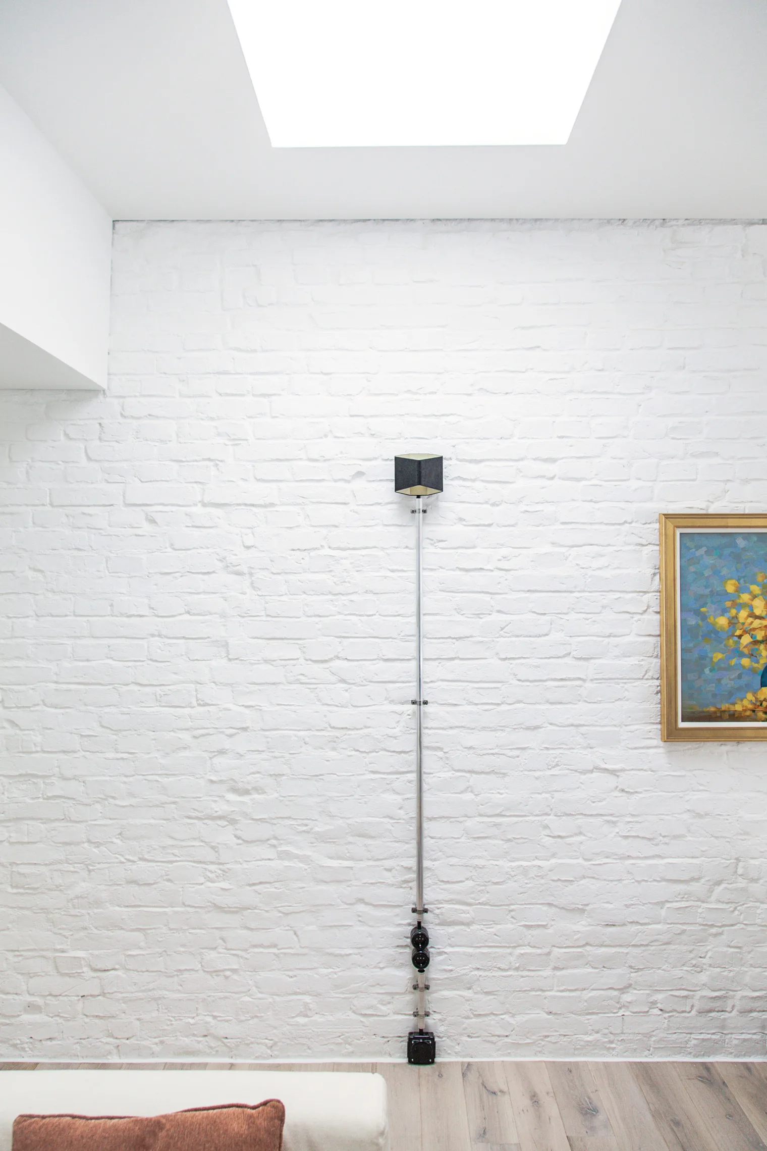 A white painted brick wall features a vertical metal conduit leading to a black cube-shaped light sconce under a bright skylight.