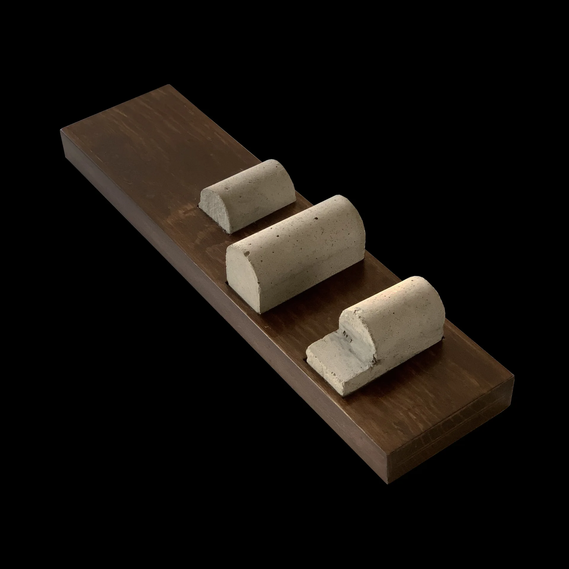 Three textured grey concrete semi-cylindrical sculptures of varying sizes are displayed on a dark brown rectangular wooden base against a black background.