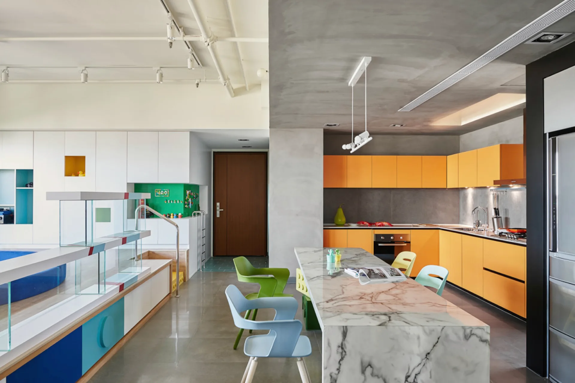 An open-plan kitchen features matte orange cabinetry, a veined marble waterfall island, and colorful molded plastic dining chairs under a split ceiling.