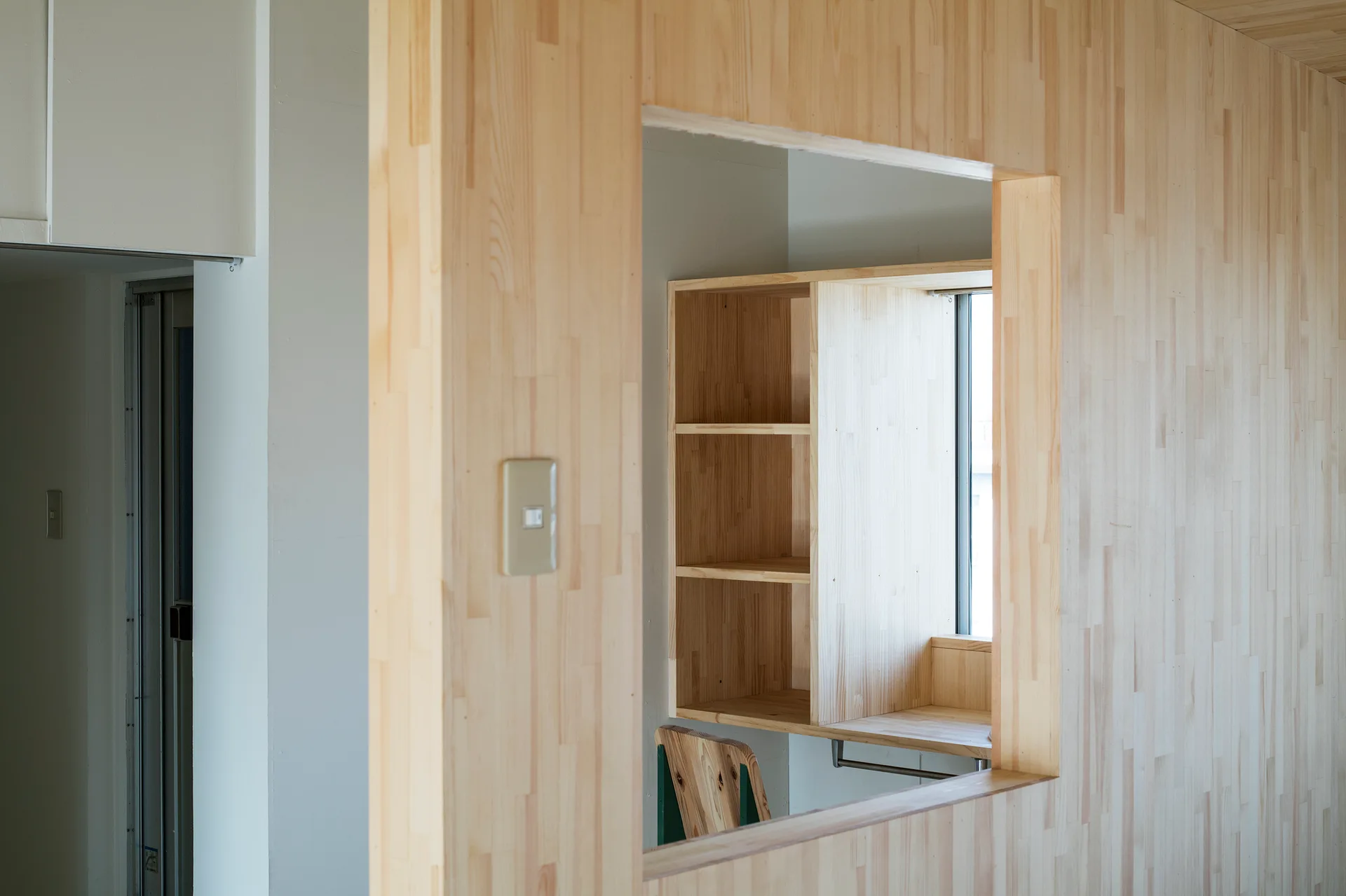 A light wood paneled partition wall features a square cutout window looking into a workspace with built-in wooden shelves.