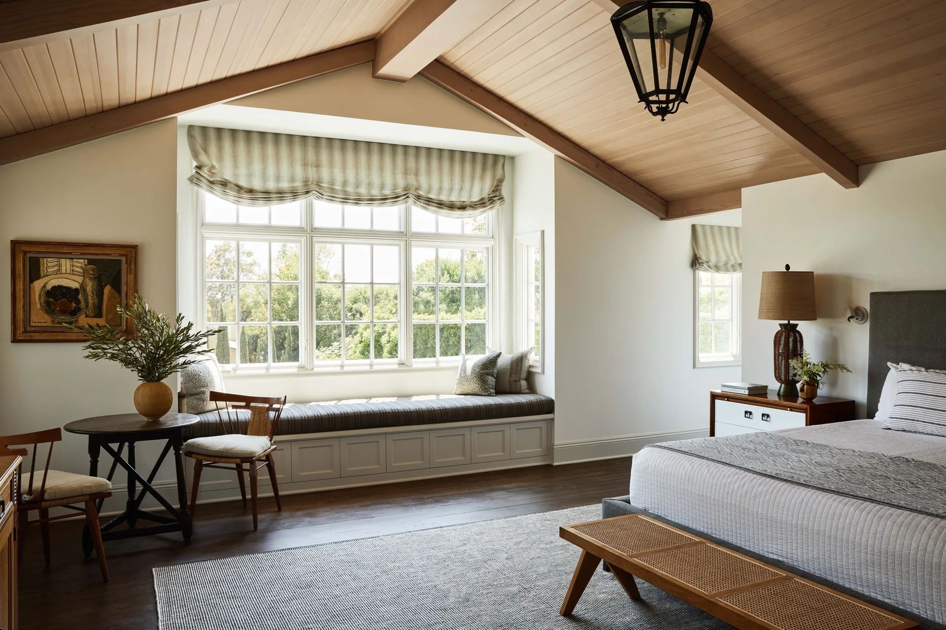 A bedroom features a vaulted wood ceiling, an upholstered bed, a woven wood bench, and a built-in window seat under a large paneled window.