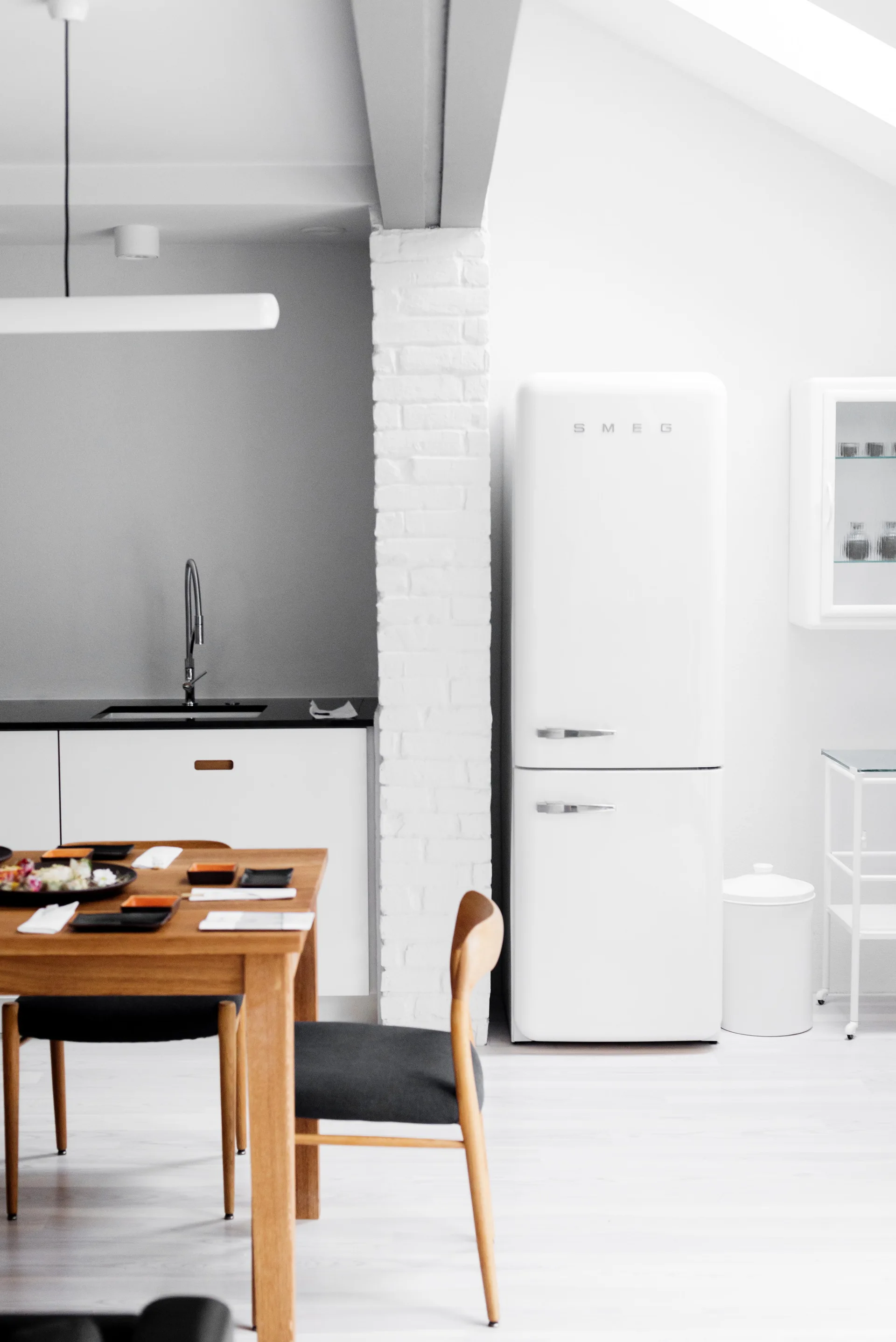 A minimalist kitchen features a retro white refrigerator beside a painted brick column and a wooden dining table with upholstered chairs.