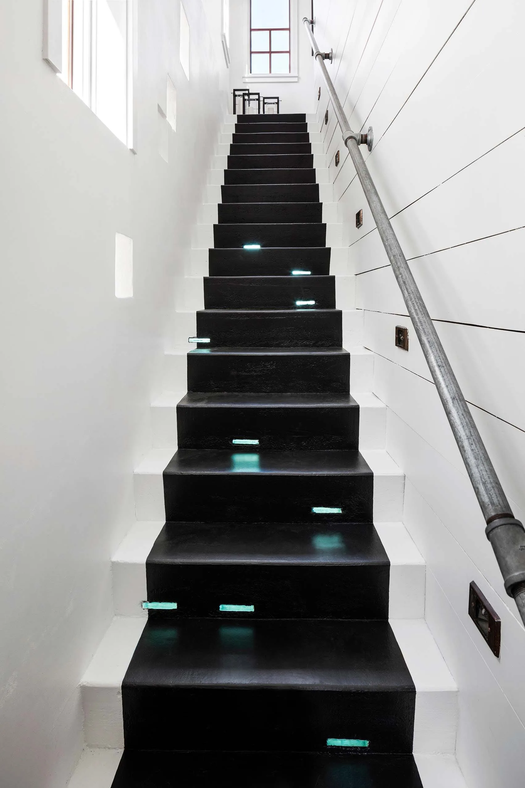 A straight black and white staircase features glowing integrated step lights, a metal pipe handrail, and white shiplap walls.