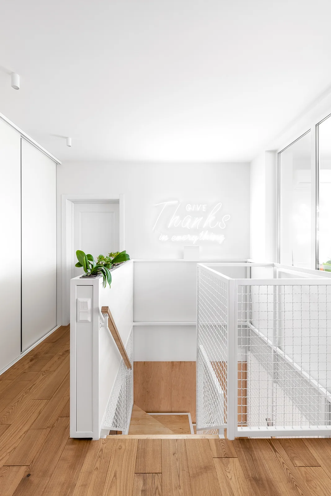 A bright minimalist stairwell featuring white walls, light wood flooring, a white metal mesh safety railing, and a glowing neon sign on the wall.