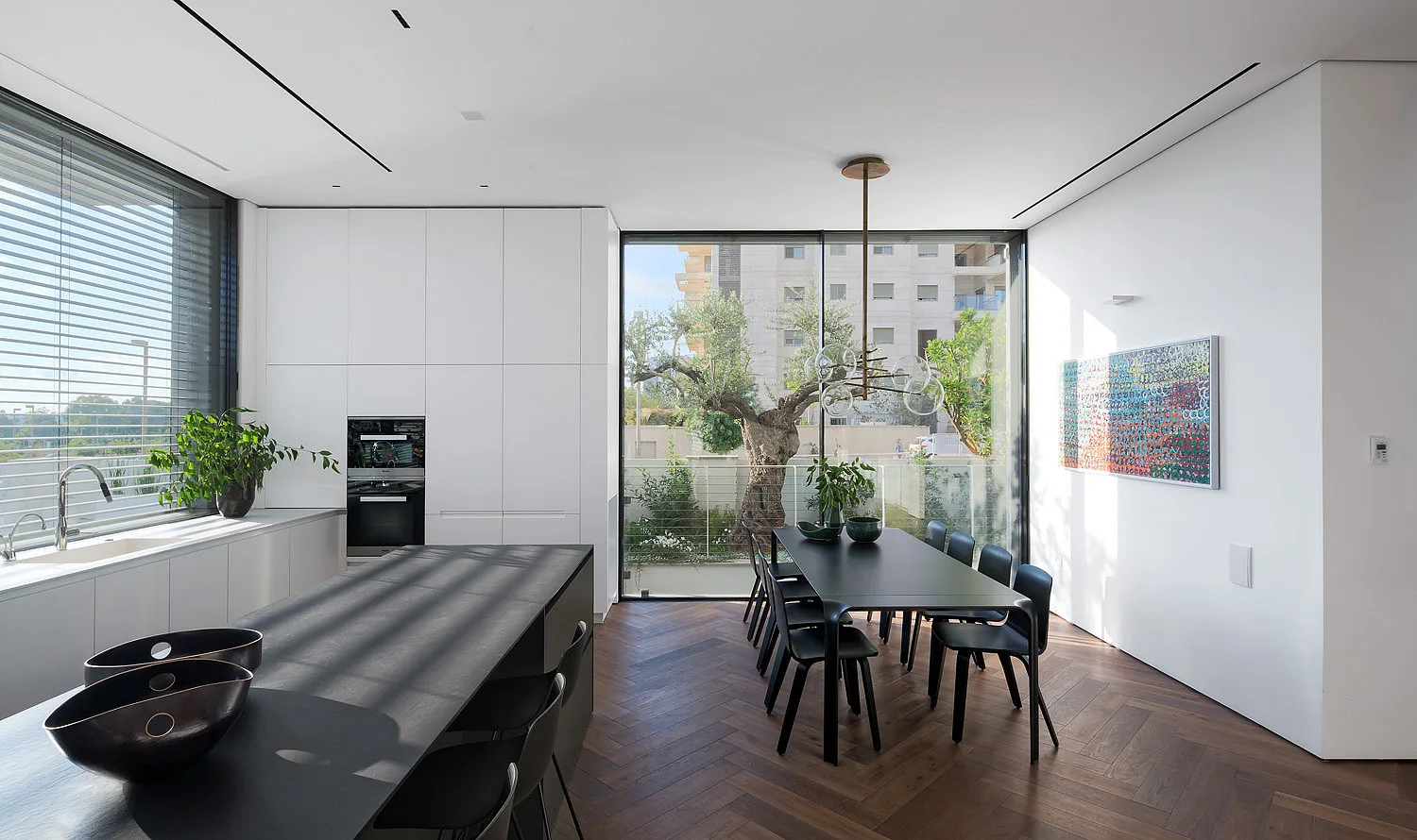 A minimalist open-plan kitchen and dining area featuring a black island, matching dining set, white cabinets, and herringbone wood floors.