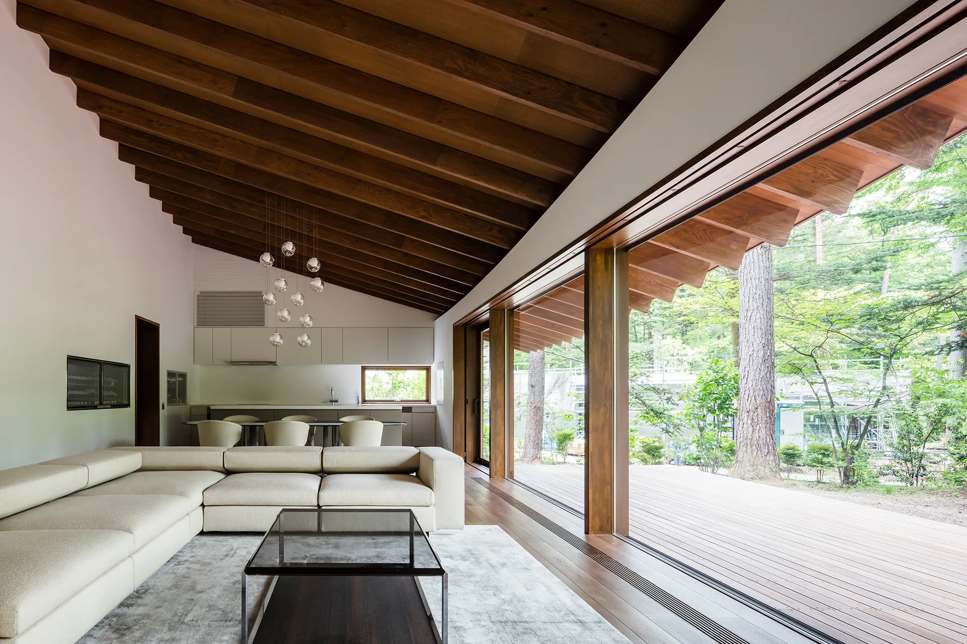 A modern living room featuring a beige sectional sofa, a sloped wood-beamed ceiling, and large glass sliding doors opening to an outdoor deck.