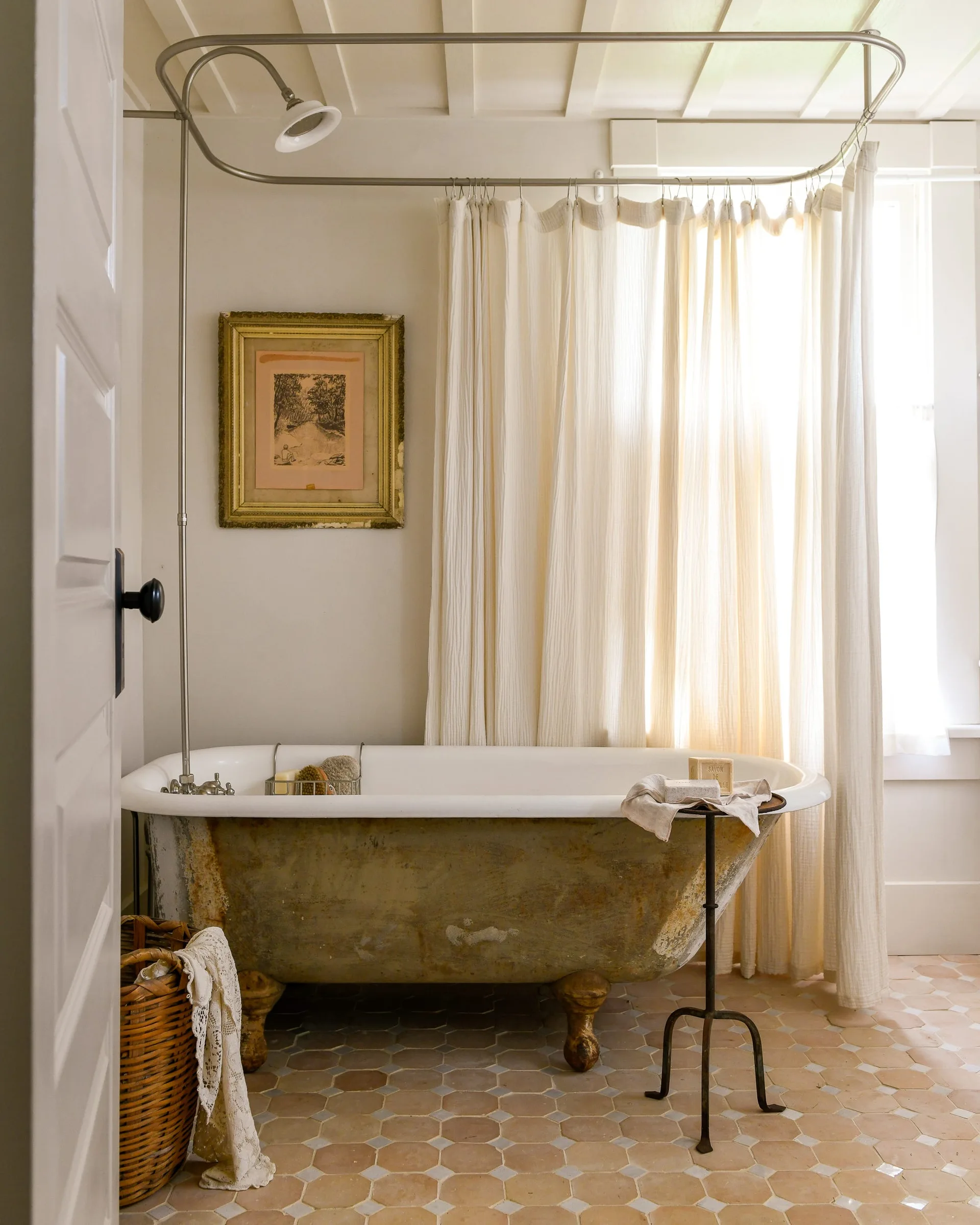 A weathered clawfoot bathtub sits on hexagonal floor tiles next to a linen shower curtain and a small metal side table.