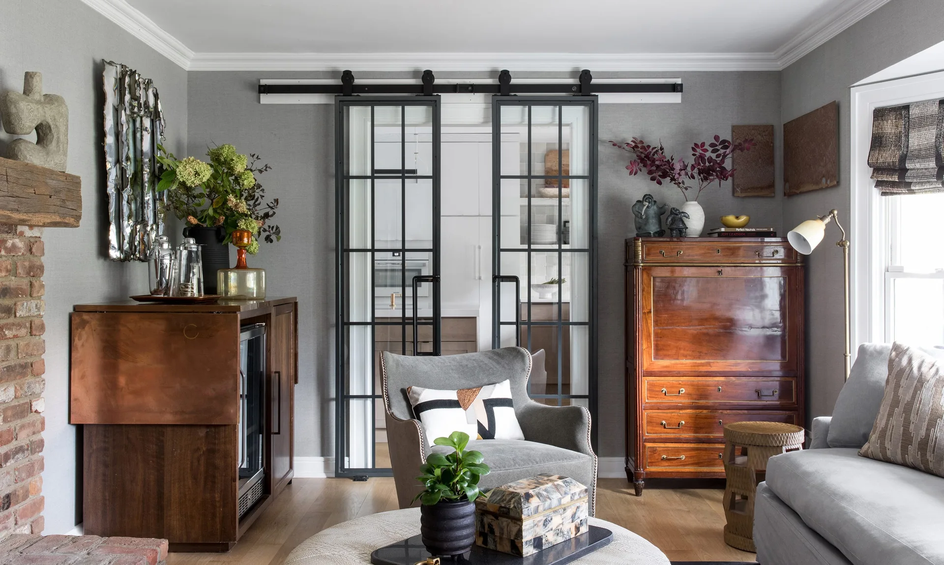 A transitional living room features a gray wingback armchair, an antique wooden secretary desk, and black-framed sliding glass doors.