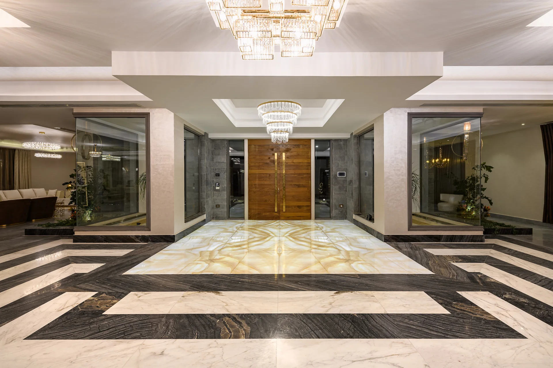 A grand entryway features a glowing backlit stone floor inlay, striped marble flooring, a large wooden double door, and tiered crystal chandeliers.