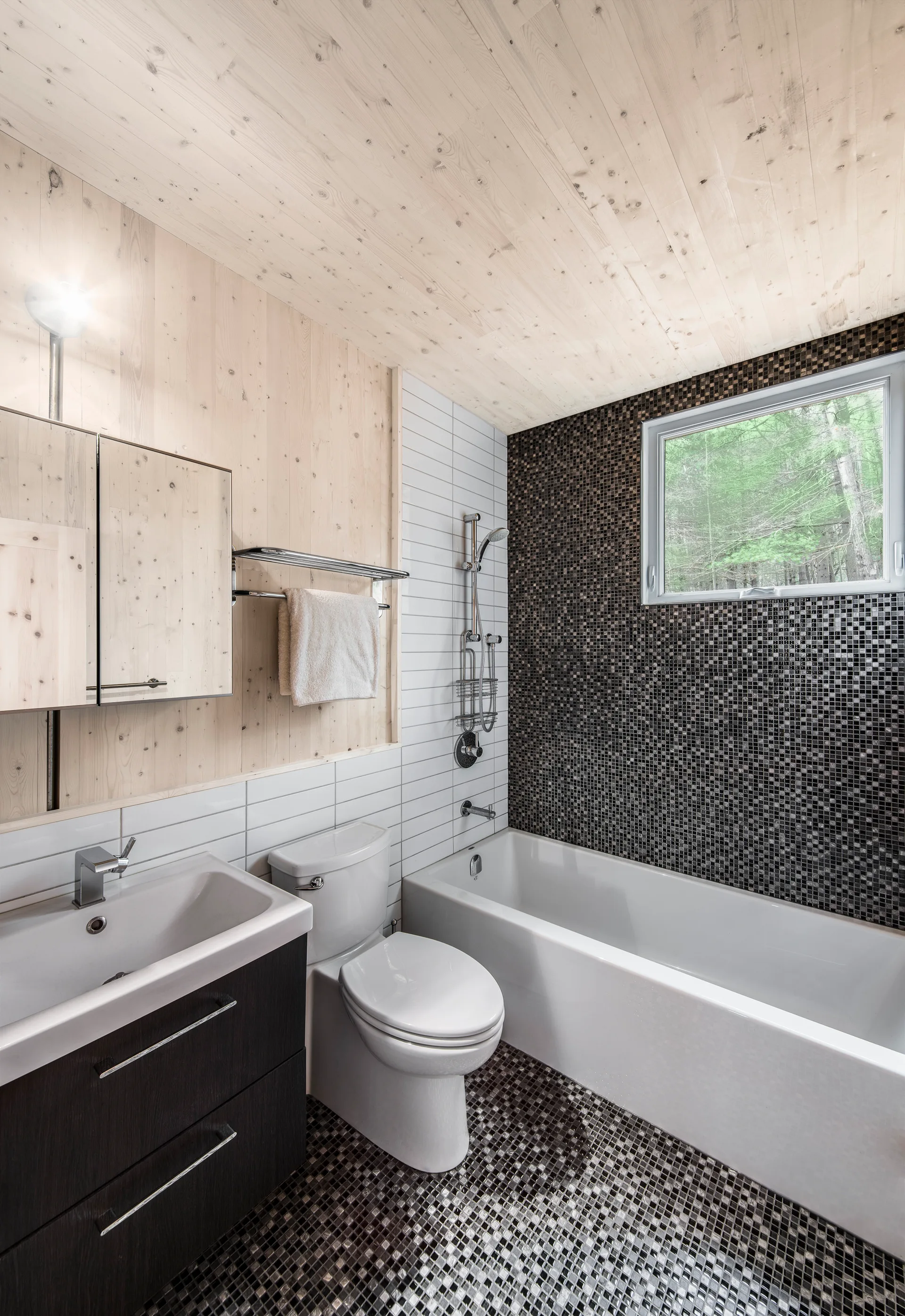 A modern bathroom featuring a white bathtub beneath a window, light wood paneled walls, white subway tile, and dark mosaic tile flooring.