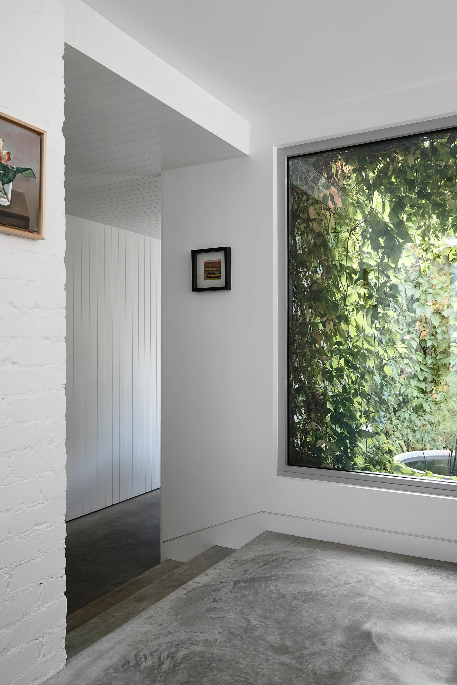 A white entryway features a polished concrete floor, vertical wall paneling, and a large window framing dense green vines outside.