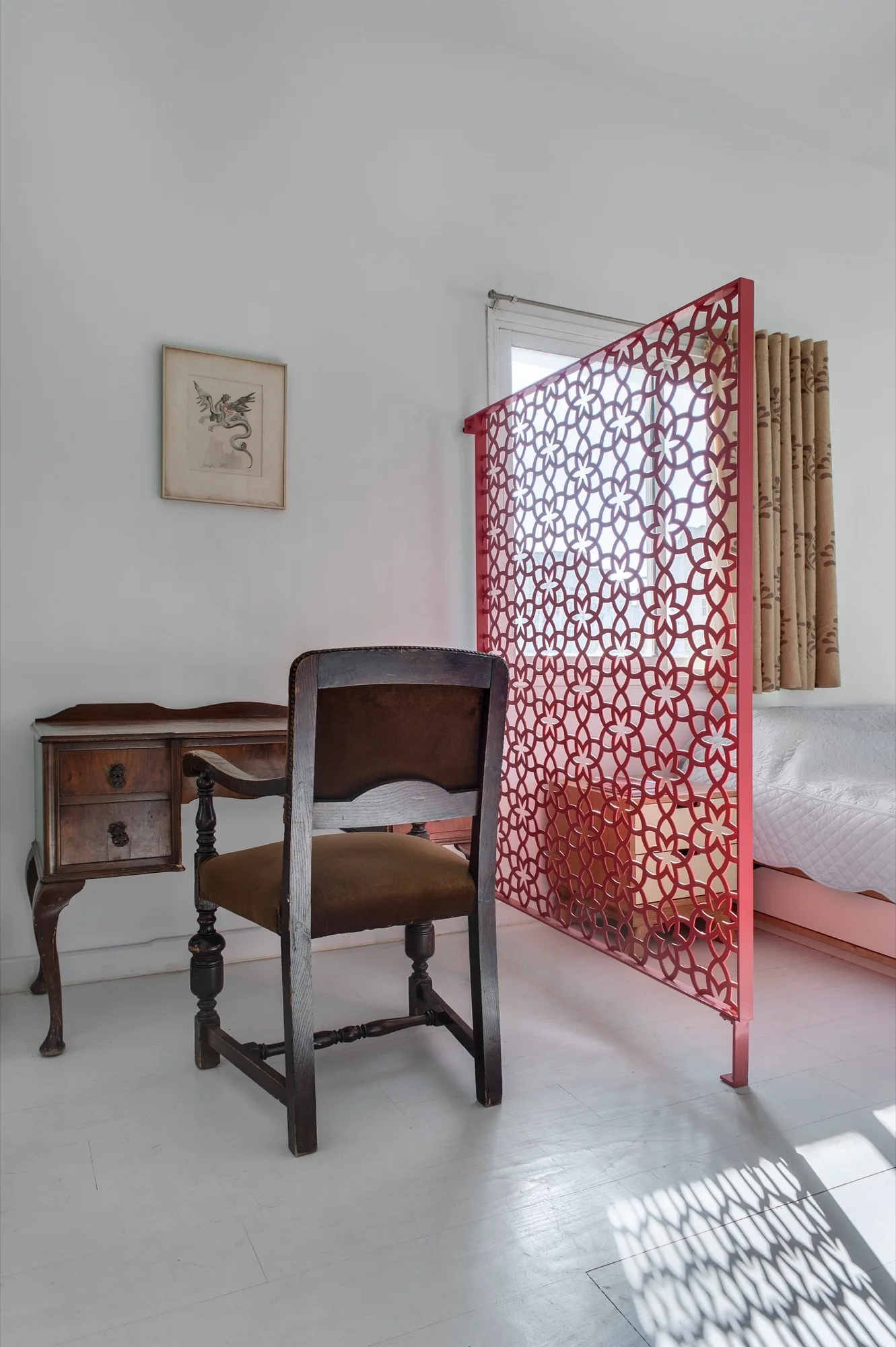 A vintage wooden desk and chair in a bedroom corner, separated from a bed by a red geometric lattice partition screen on white wood floors.