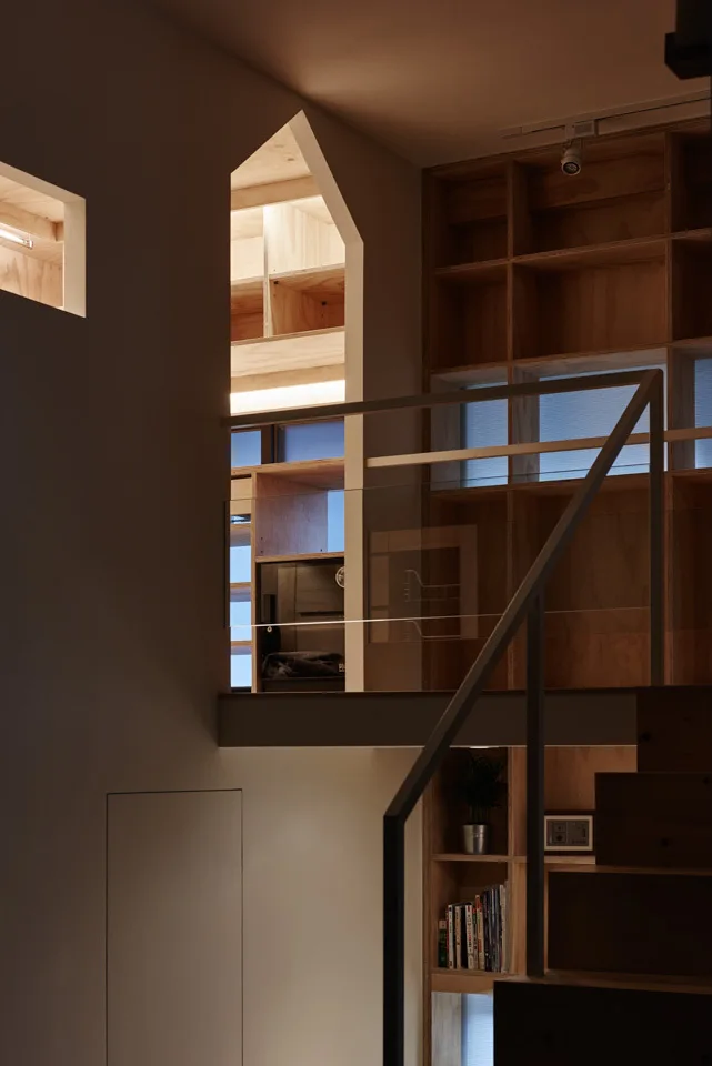 A modern staircase features a glass safety railing, a pointed archway cutout, and large built-in plywood grid shelving with warm integrated lighting.