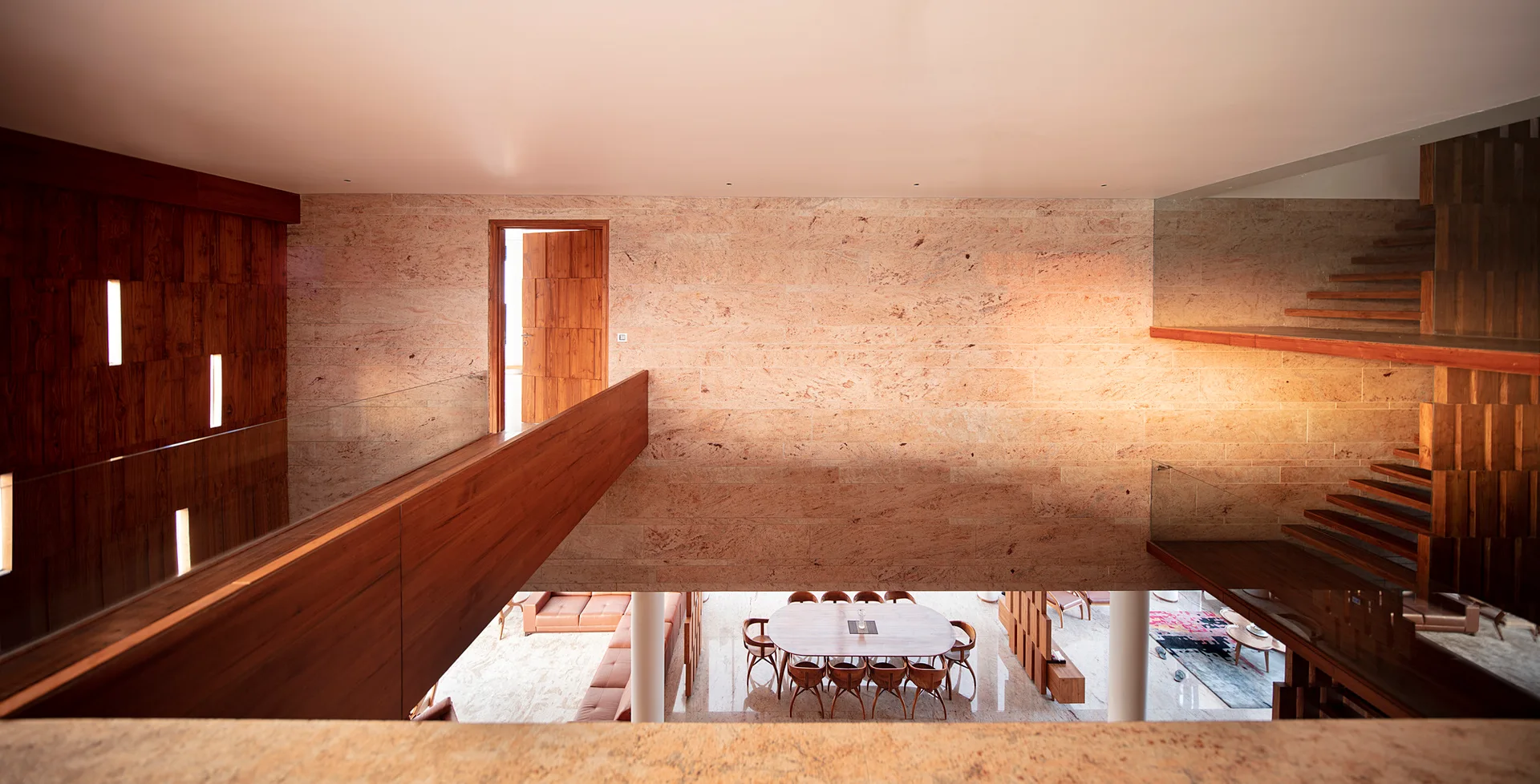A view from a wood-paneled mezzanine looks down at an open-concept dining area, featuring a large textured stone wall and a floating wooden staircase.