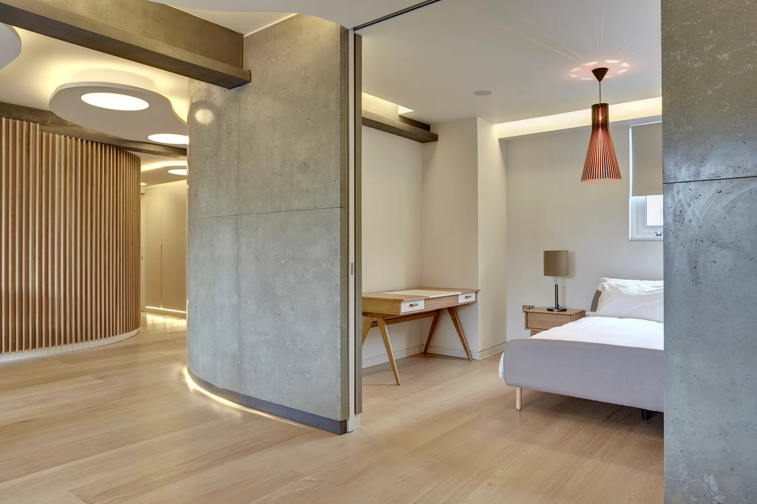 A modern bedroom features a grey curved concrete partition wall, light wood flooring, a wooden writing desk, and a bed with a hanging pendant light.