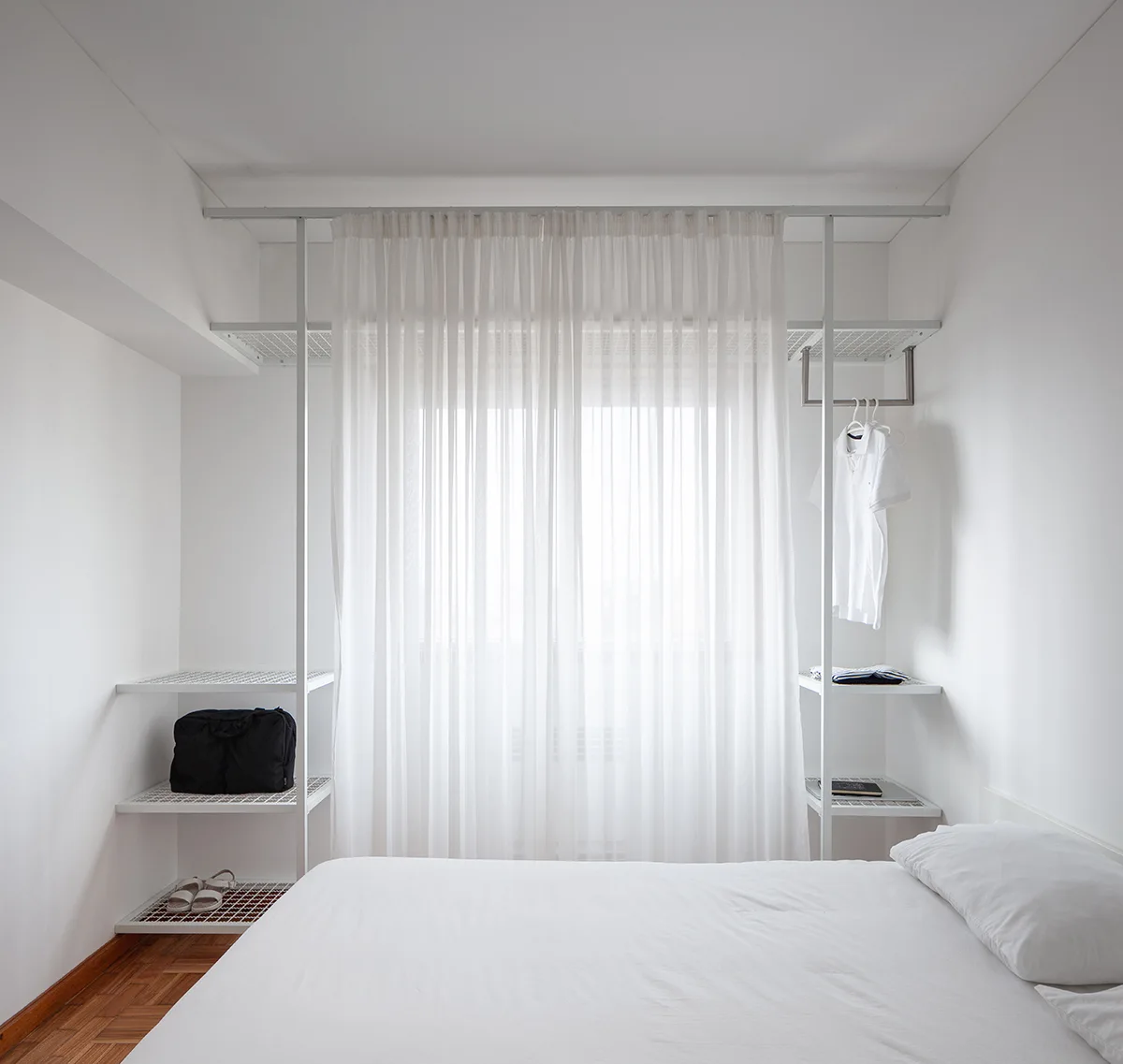 A minimalist bedroom features a white bed, wood parquet flooring, and a white metal open wardrobe shelving unit surrounding a window with sheer curtains.