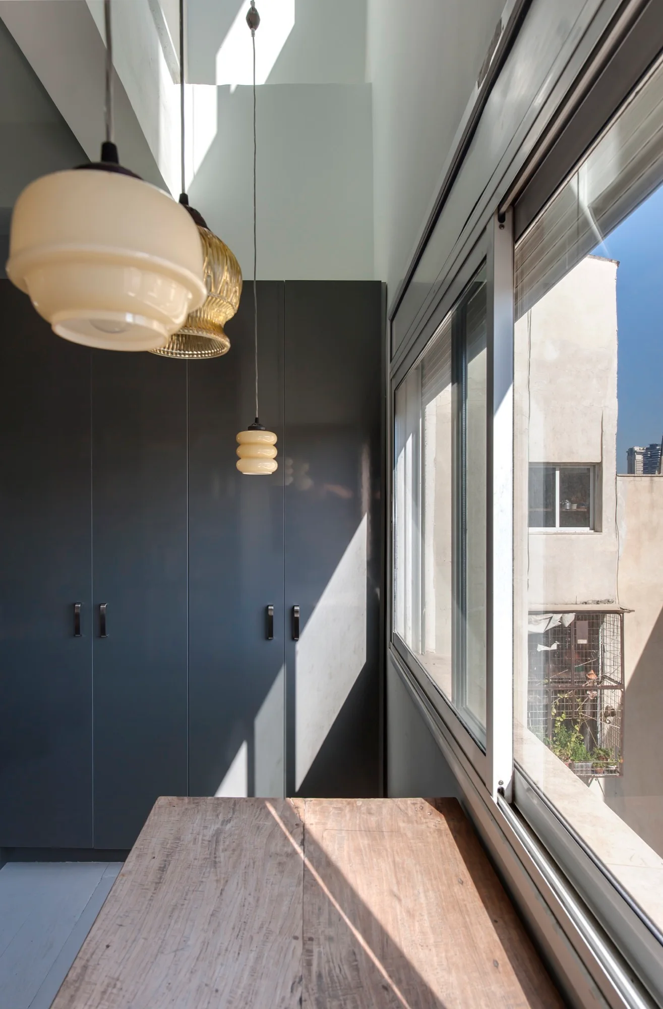 A bright interior space features matte grey storage cabinets, a wooden tabletop, and retro glass pendant lights hanging next to a large window.