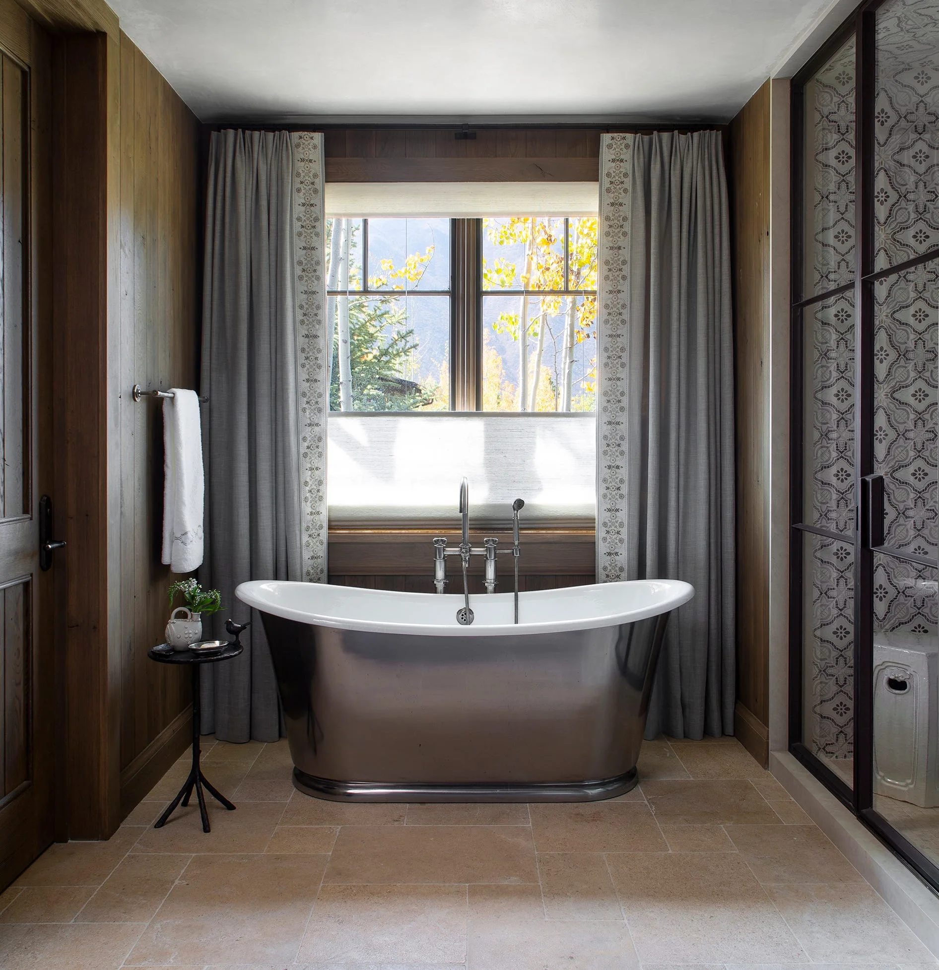 A metallic freestanding bathtub is centered beneath a window with grey curtains in a bathroom featuring wood-paneled walls and a stone tile floor.