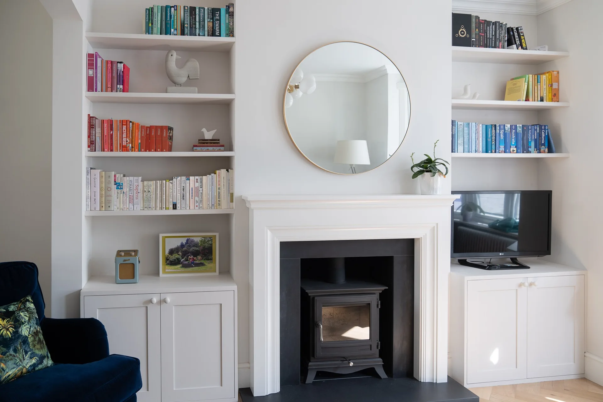 A living room featuring white built-in bookshelves flanking a fireplace with a wood stove, a gold round mirror, and a blue armchair.