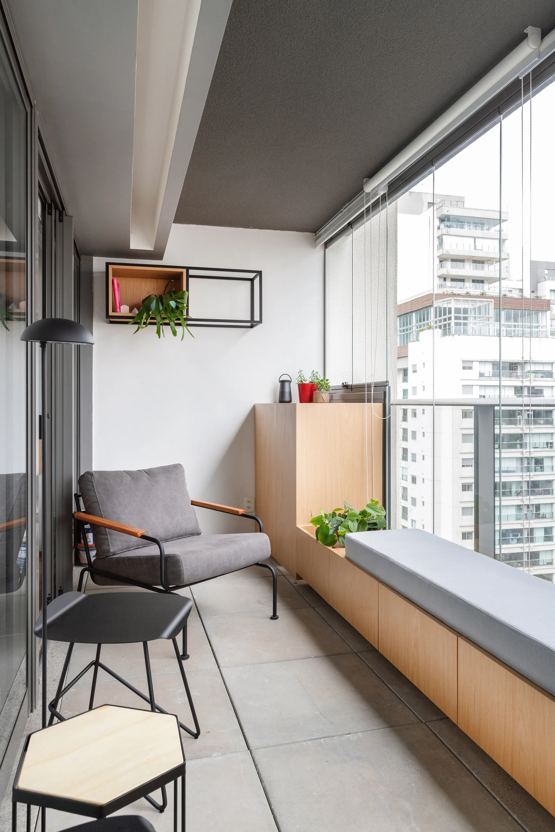A modern enclosed balcony features a light wood built-in bench with a grey cushion, a grey armchair, and glass walls overlooking city buildings.