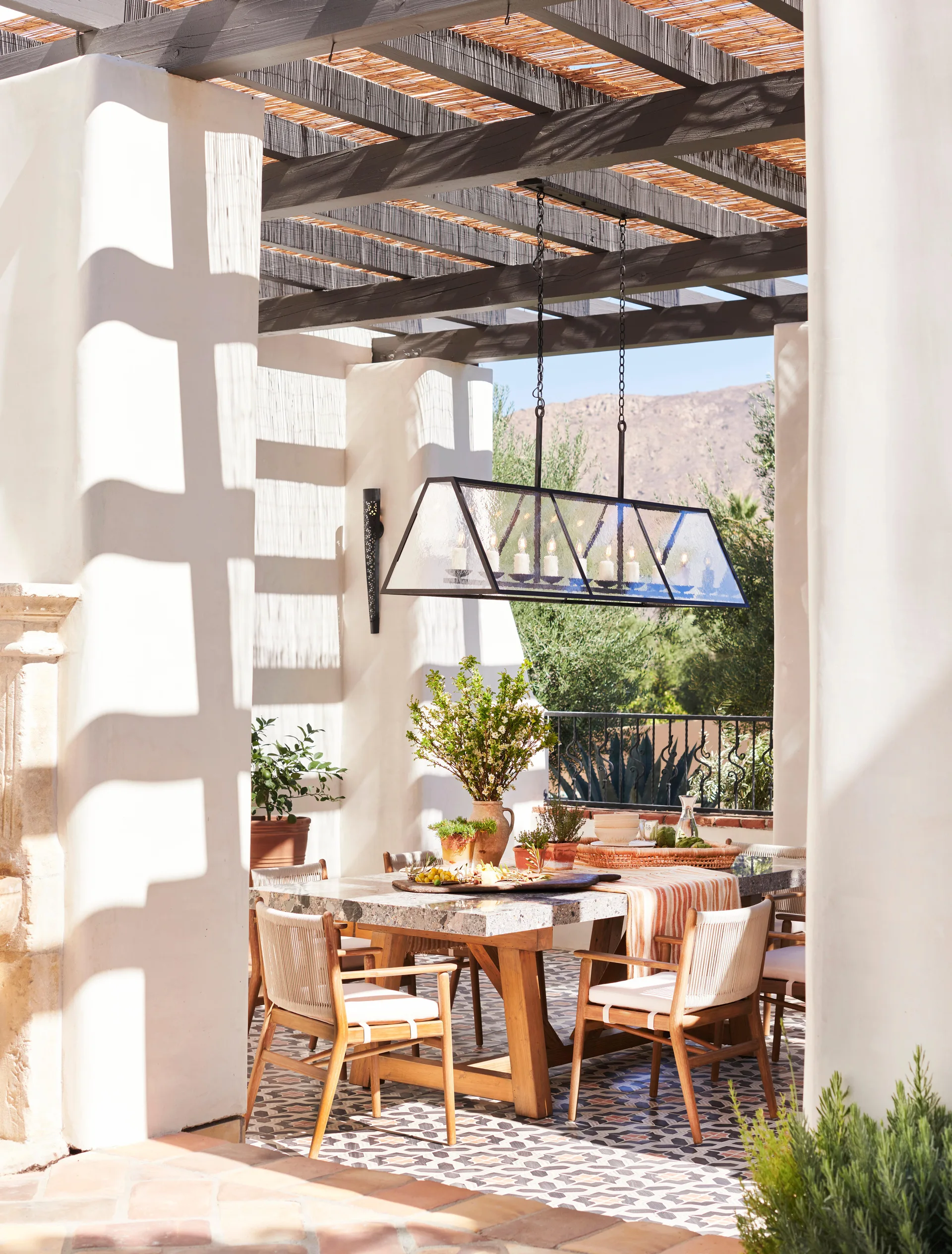 An outdoor patio dining space features a stone-topped table with wooden chairs under a timber pergola, set on a patterned tile floor.