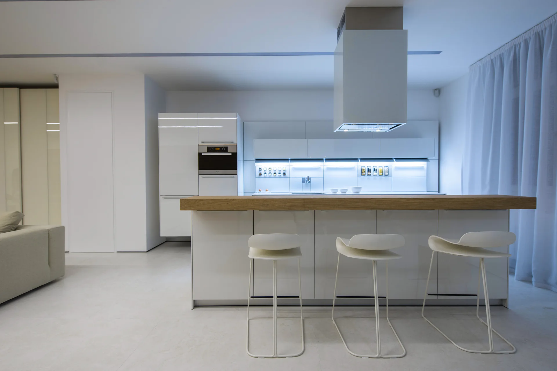 Minimalist white kitchen featuring an island with a thick wood countertop, three bar stools, and glossy cabinets with integrated lighting.