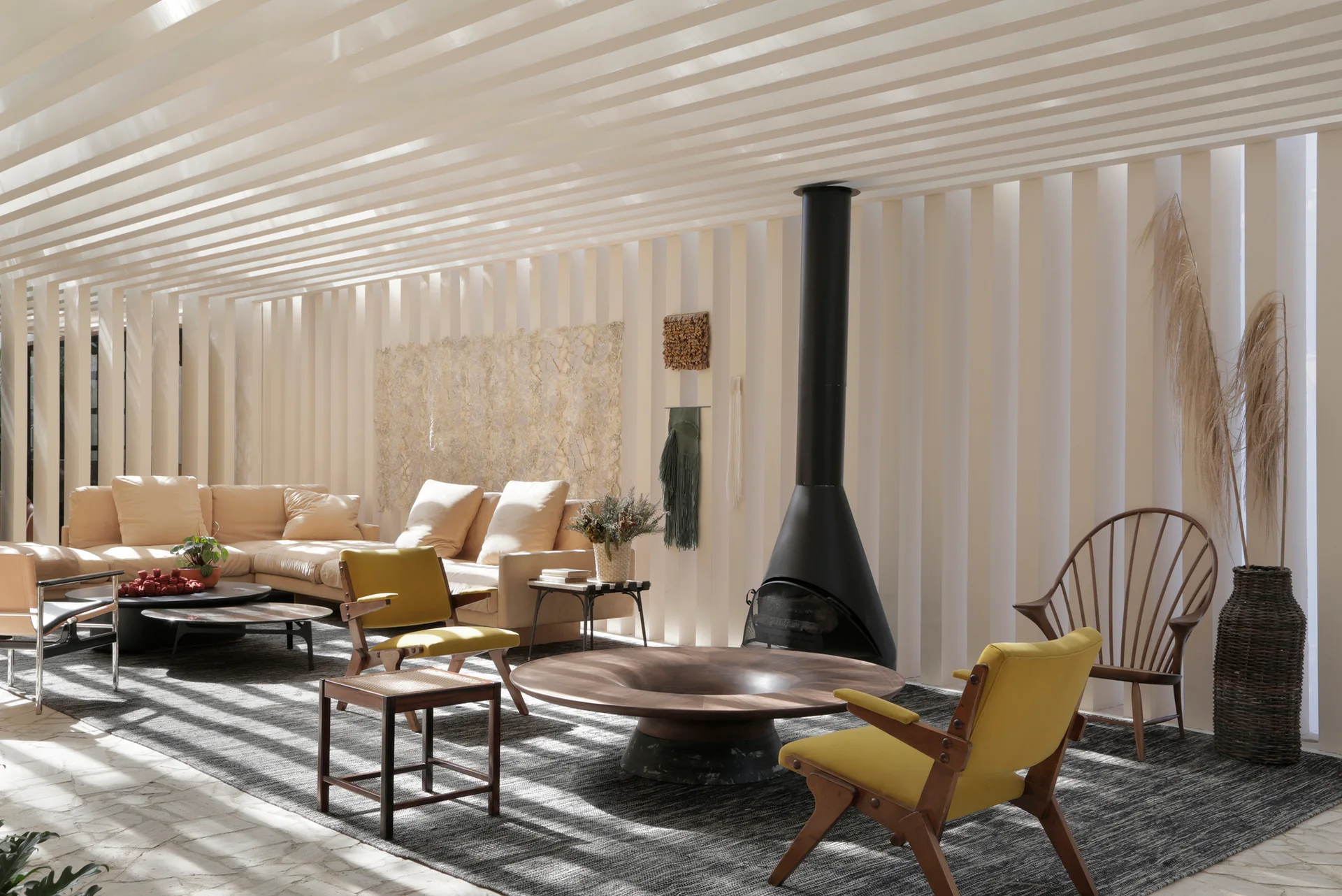 A living room with vertical slatted walls and ceiling, a black conical fireplace, two yellow armchairs, and a low wood coffee table.