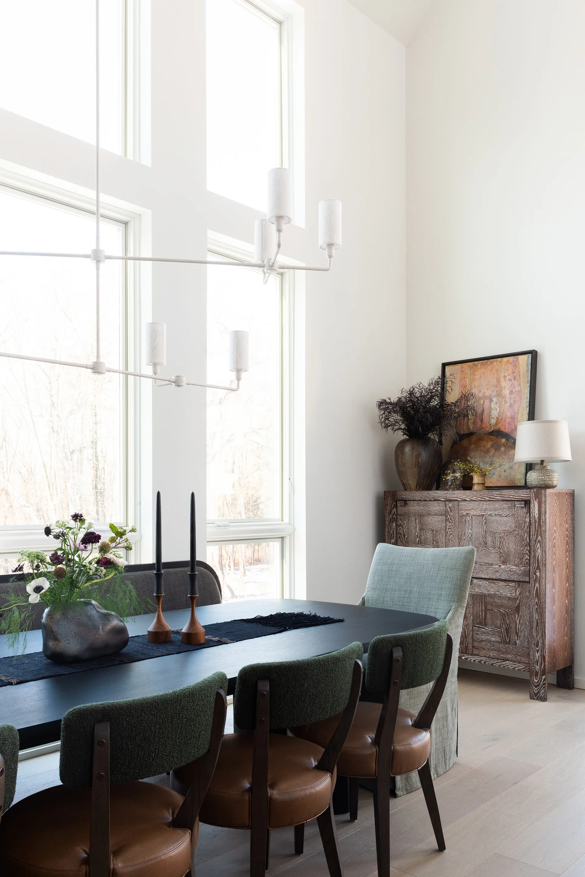 A bright dining room features a black table with green and brown chairs, set against tall windows next to a textured wooden cabinet.